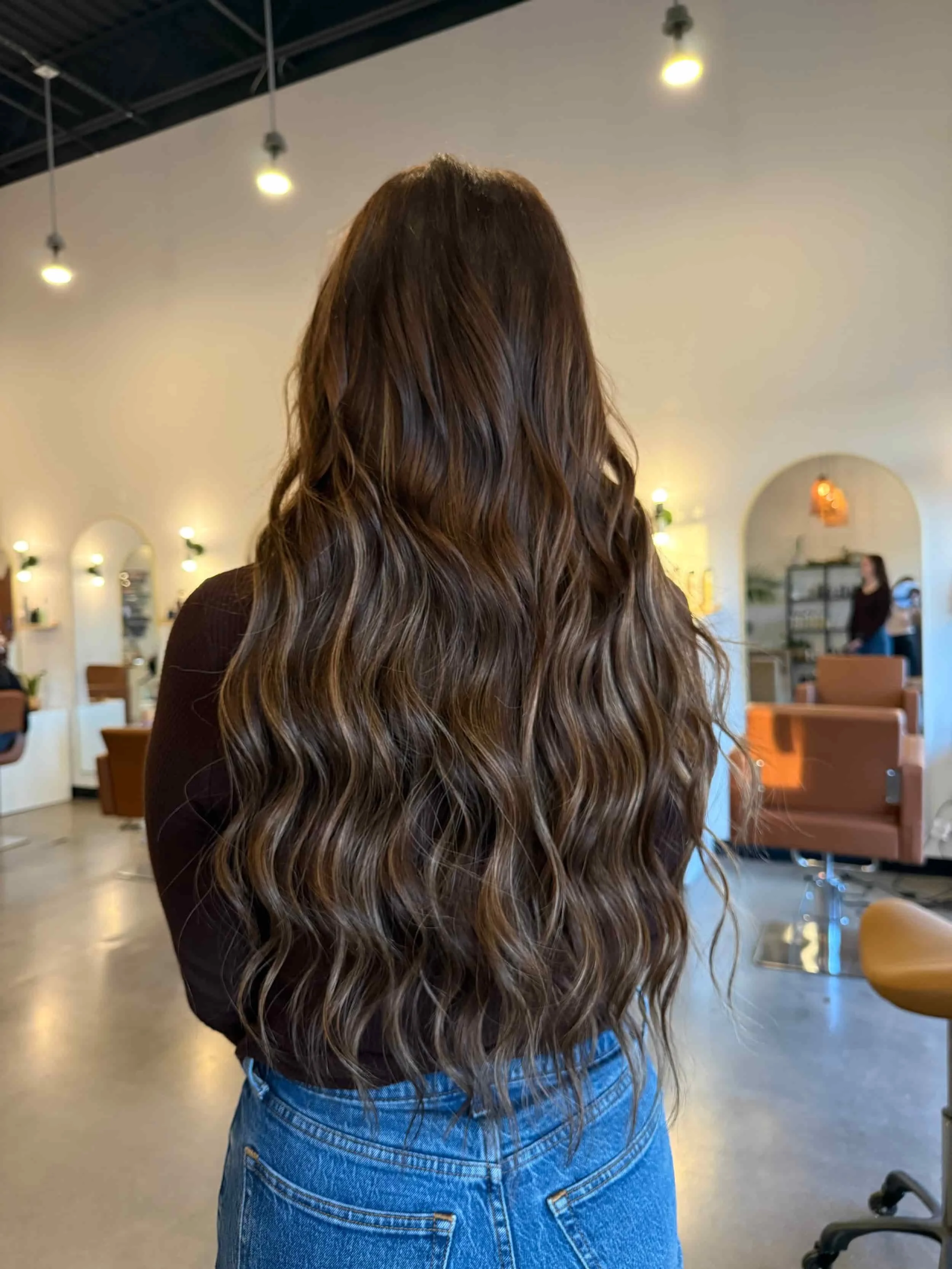 Back view of a woman with very long, rich dark brown hair featuring subtle bronze and caramel balayage highlights and new tape-in extensions styled in defined beach waves, shown in a bright open hair salon