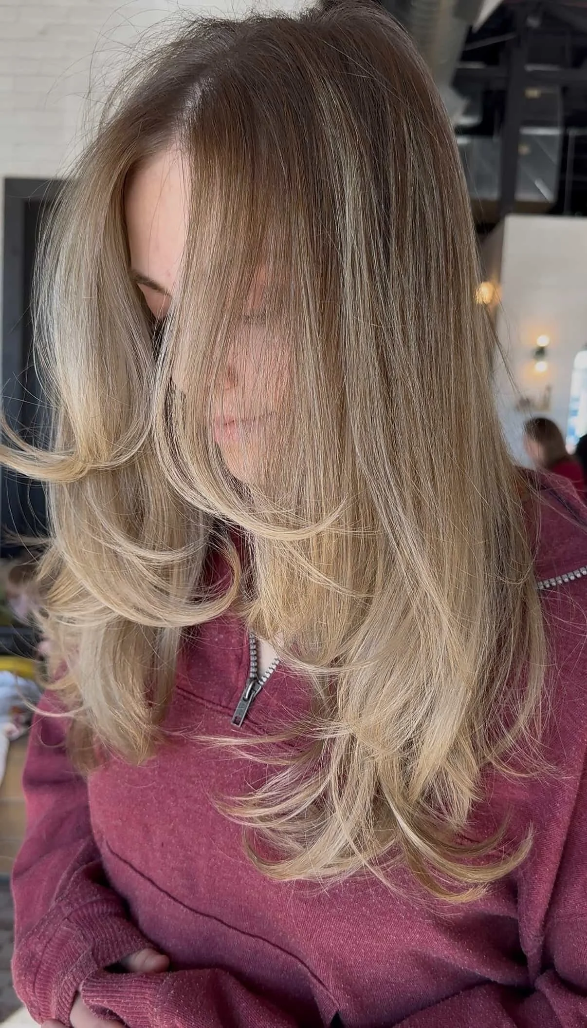 Close-up of a woman with medium-length dark blonde hair featuring lighter blonde balayage highlights and a layered blow-dry finish, with her face obscured by her hair as she looks down in a salon setting.