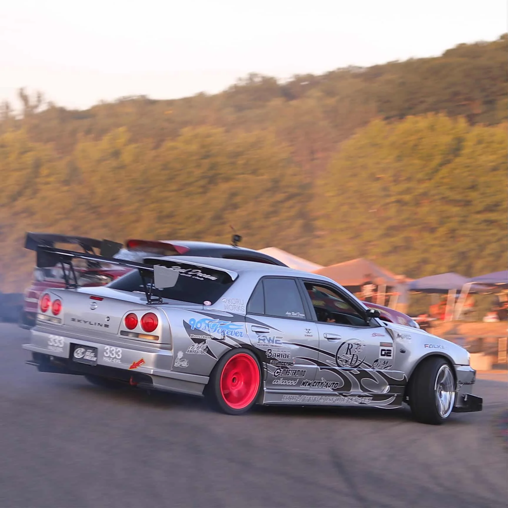 A silver race car with red wheels drifting on a track during a motorsport event, with a background of mountains and tents.