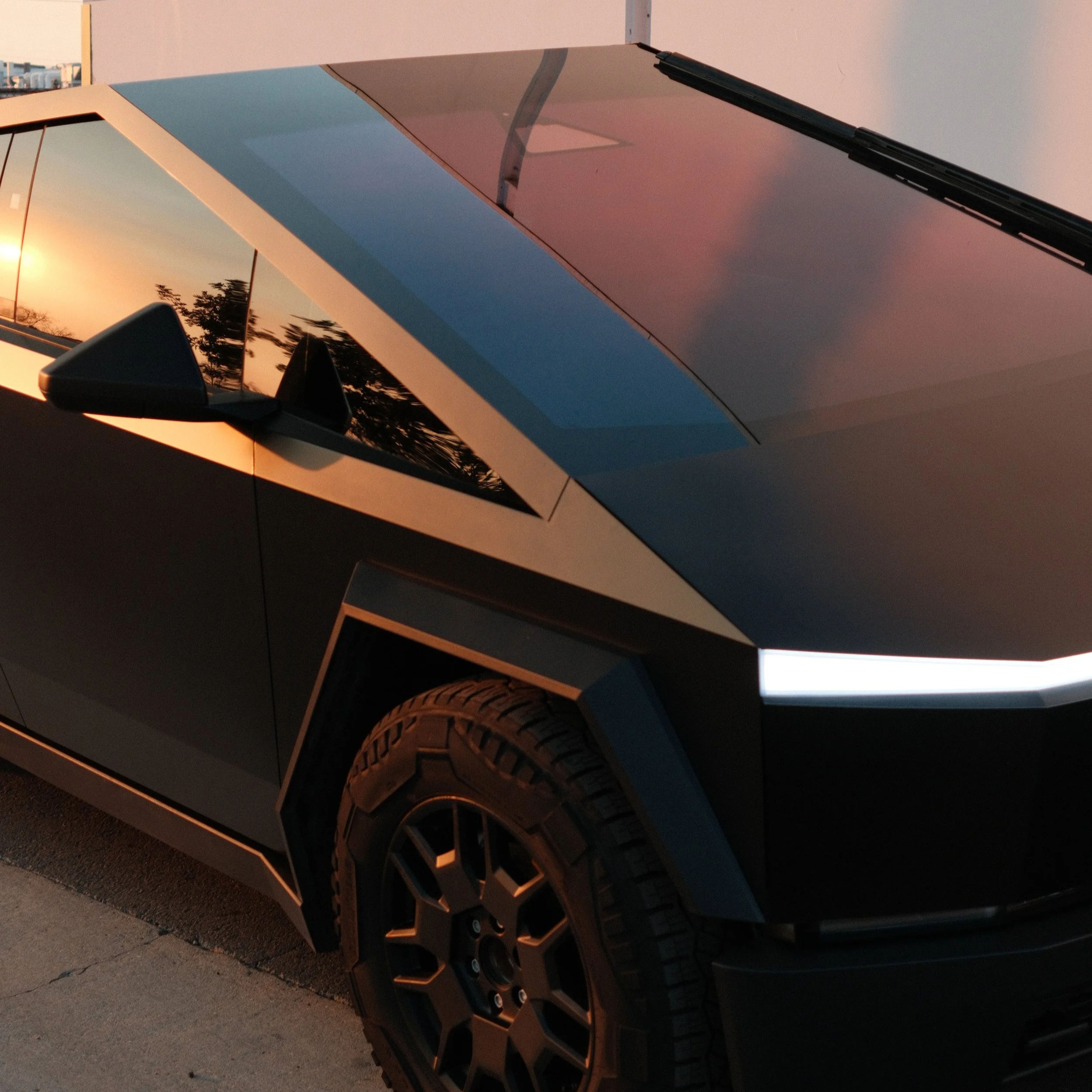 Close-up of a black vehicle, possibly an electric truck, with a modern angular design, reflecting sunset colors on its shiny surface, showing details of the tire, wheel, and part of the front headlight.