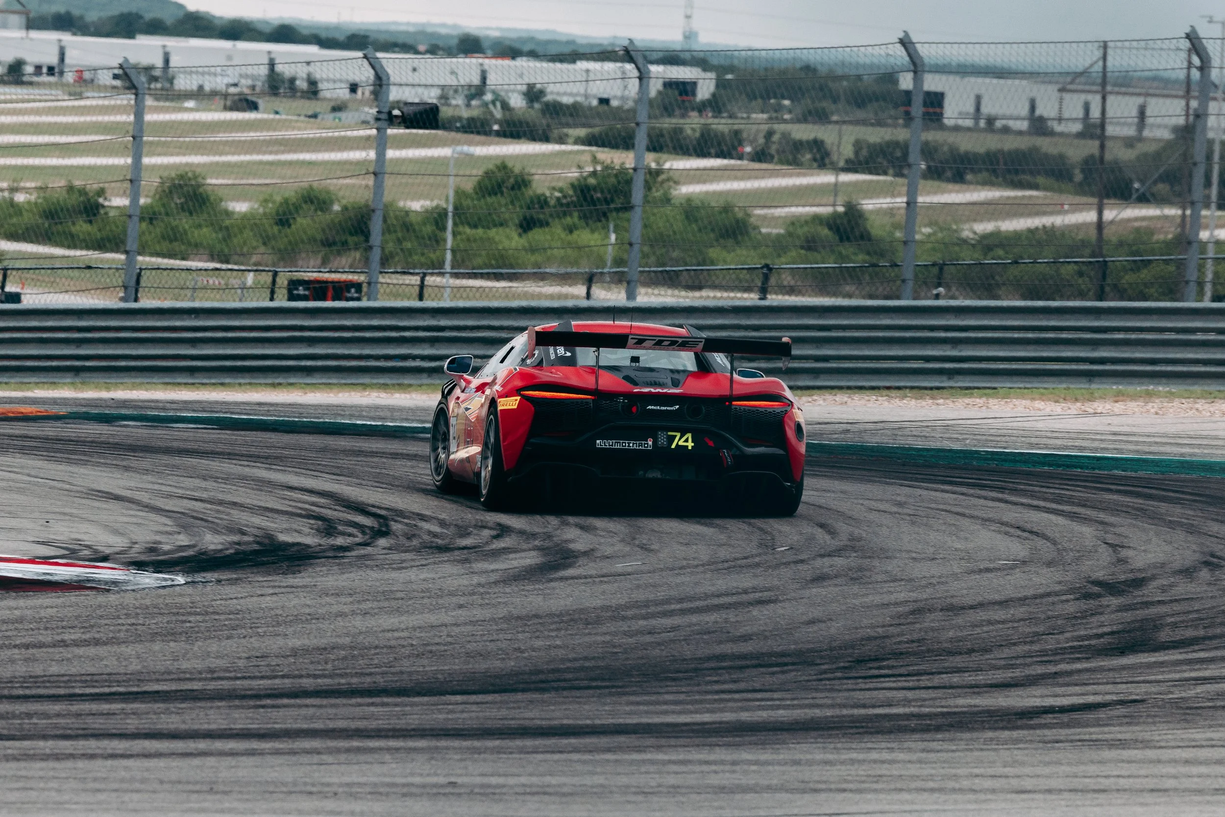 A red race car speeding on a race track with tire marks, surrounded by barriers and fences, during daytime.
