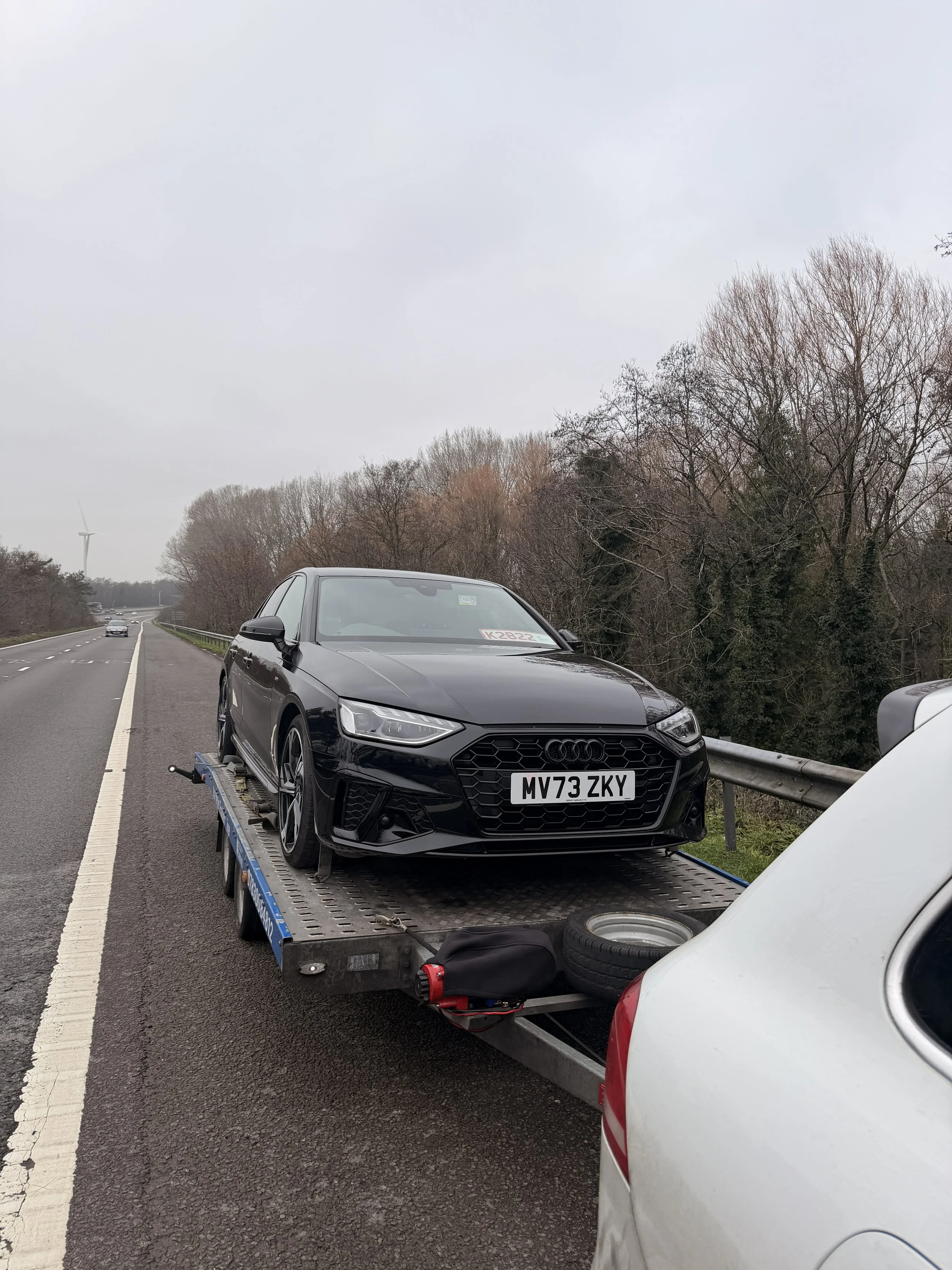 A black Audi sedan on a flatbed tow truck on the side of a highway, with a white vehicle partially visible in the foreground