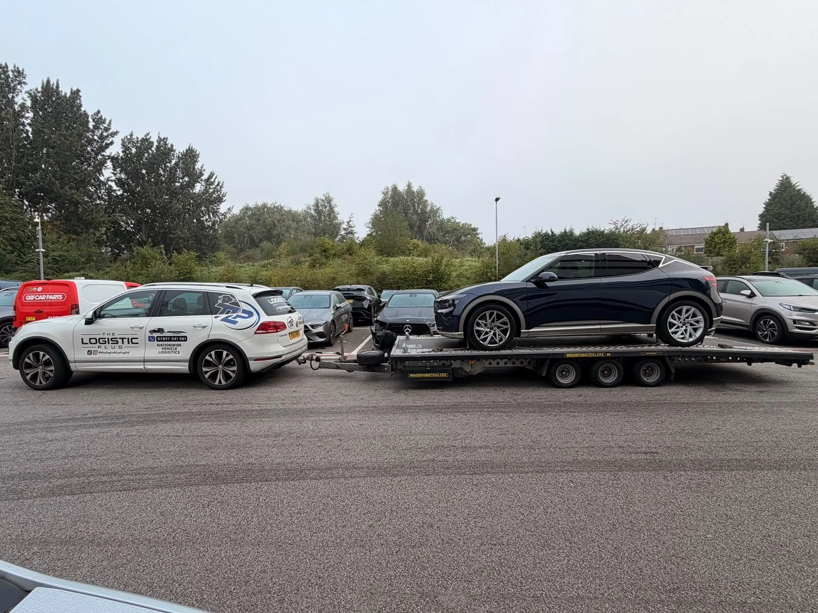 A black Mercedes-Benz SUV on a flatbed truck in a parking lot with other parked cars and greenery in the background.