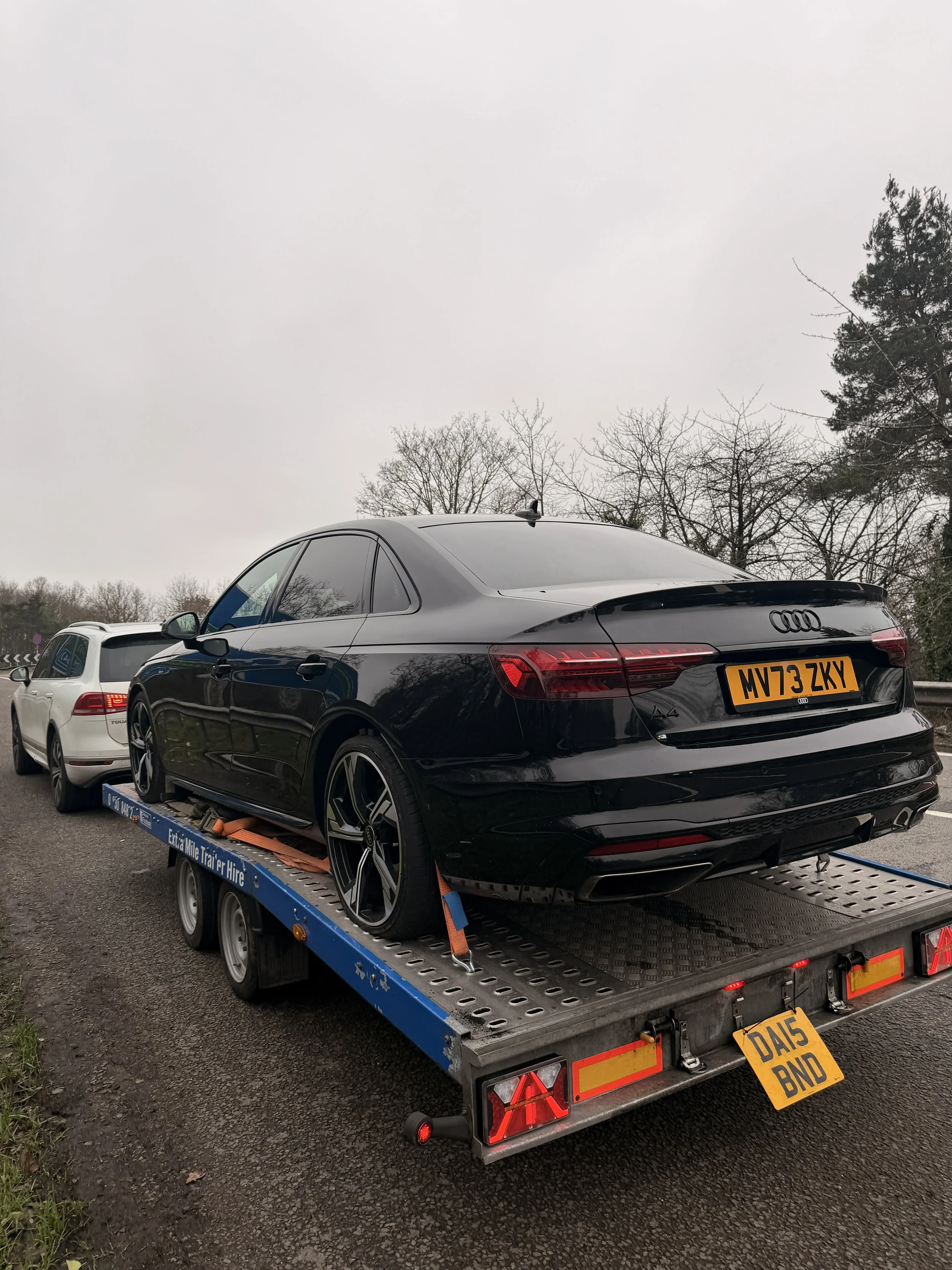 A black Audi car loaded on a flatbed tow truck, with a white vehicle behind it, on an overcast day with leafless trees in the background.