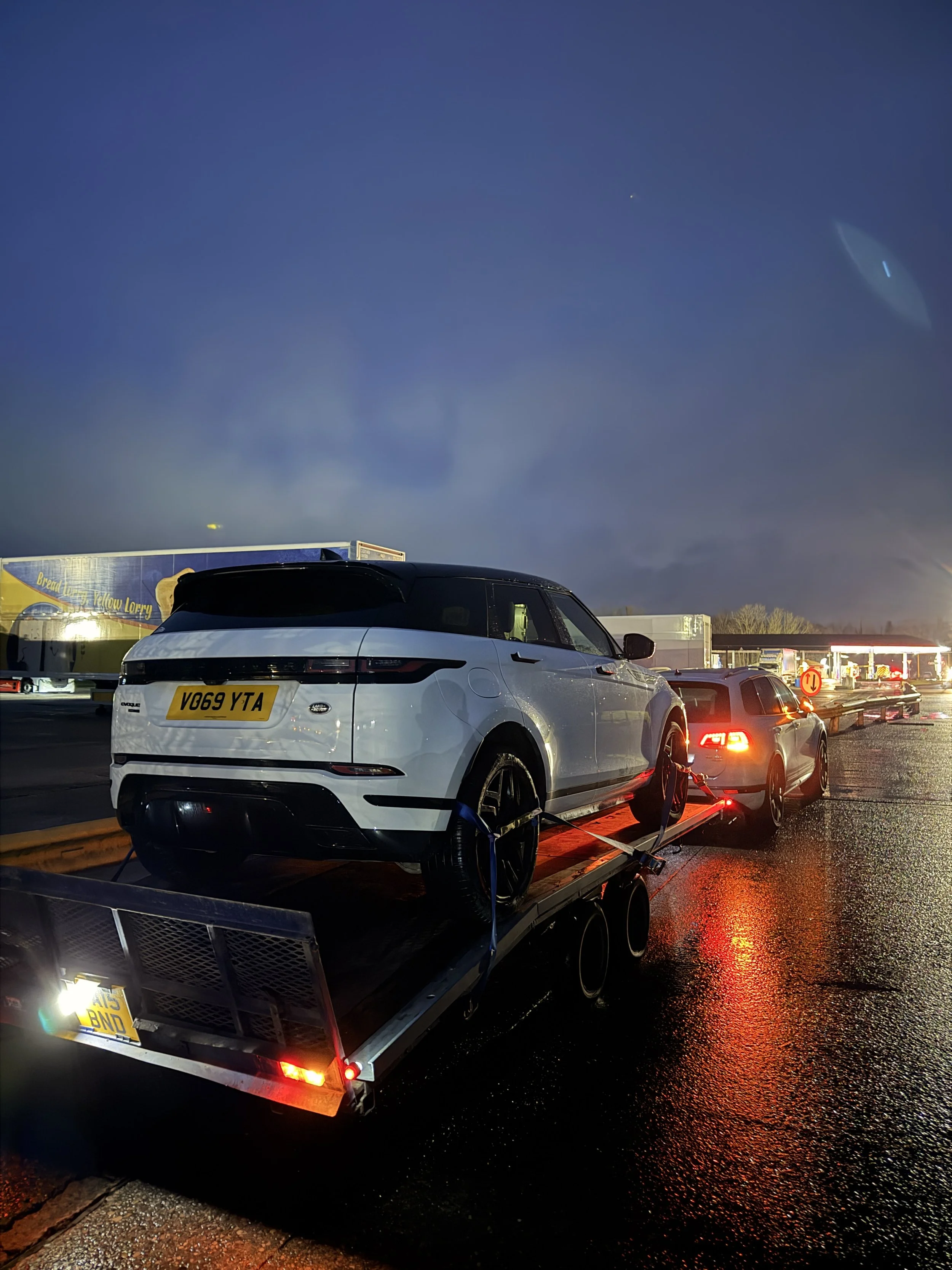 A white and black Range Rover Velar with a UK license plate, parked on a wet street at dusk, attached to a trailer carrying a black sports car. The scene has a cloudy sky with a light reflecting on the wet pavement and a truck visible in the backgrou