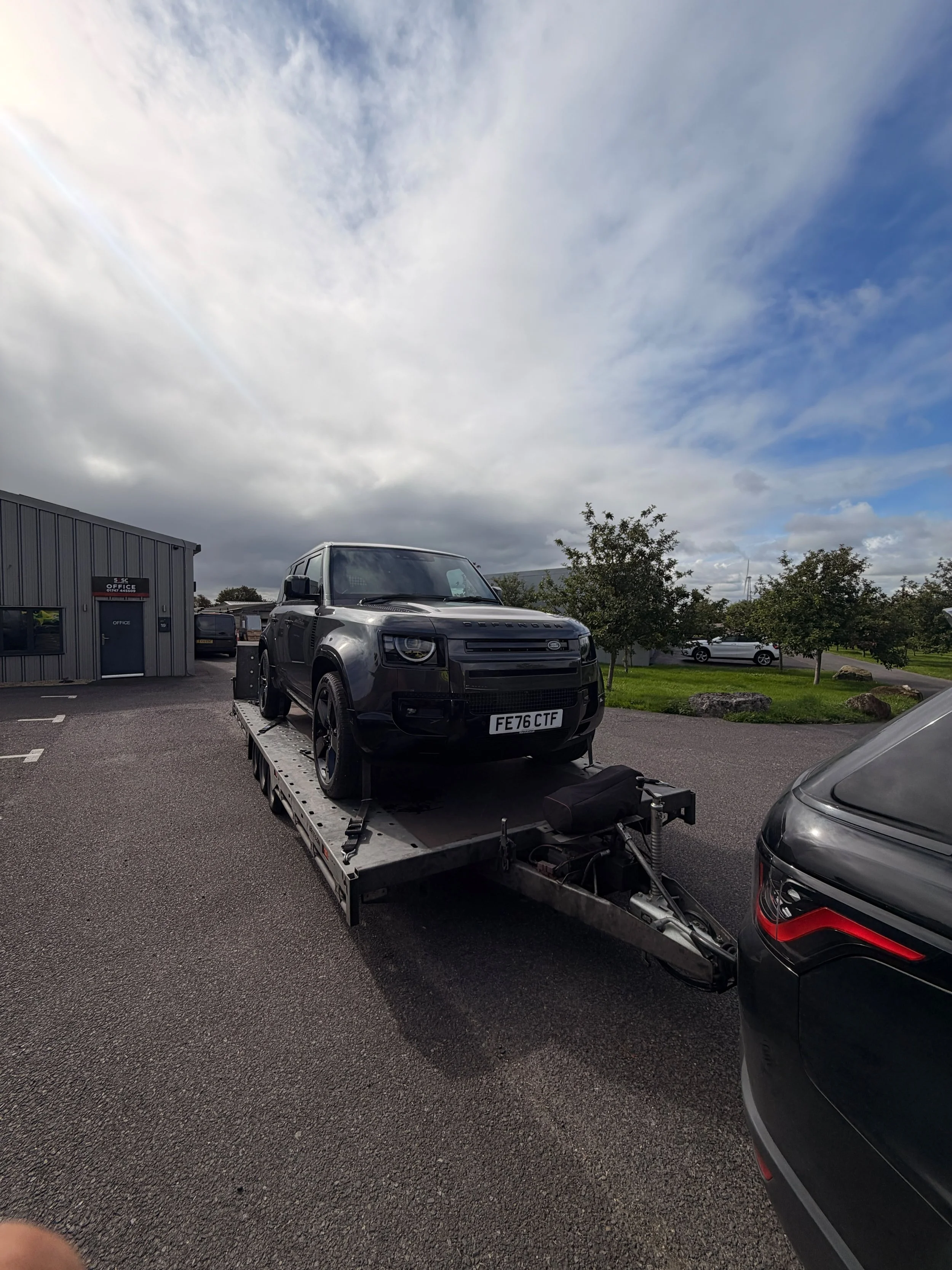 A black Land Rover vehicle on a flatbed tow truck in a parking lot on a cloudy day.