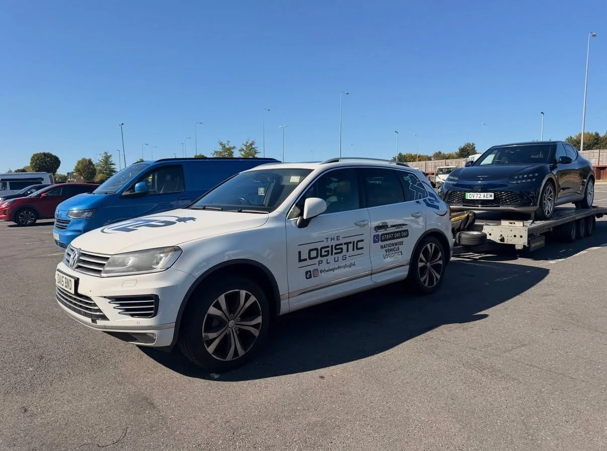 White vehicle with 'The Logistic Plus' branding towing a black car on a flatbed in a parking lot under a clear blue sky.