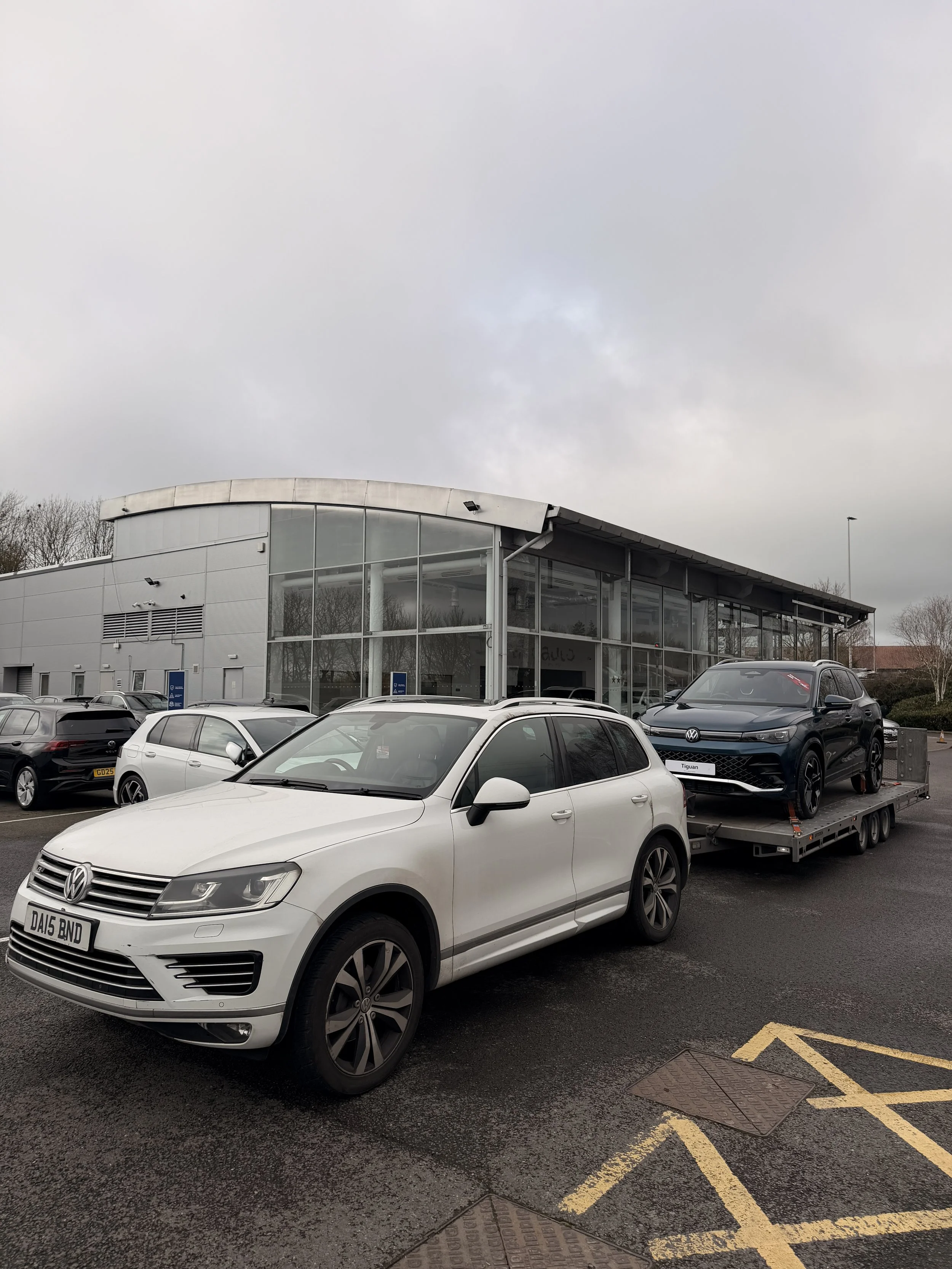A white Volkswagen sedan parked in a parking lot in front of a modern glass building, with a black Volkswagen Tiguan on a trailer behind it, under a cloudy sky.