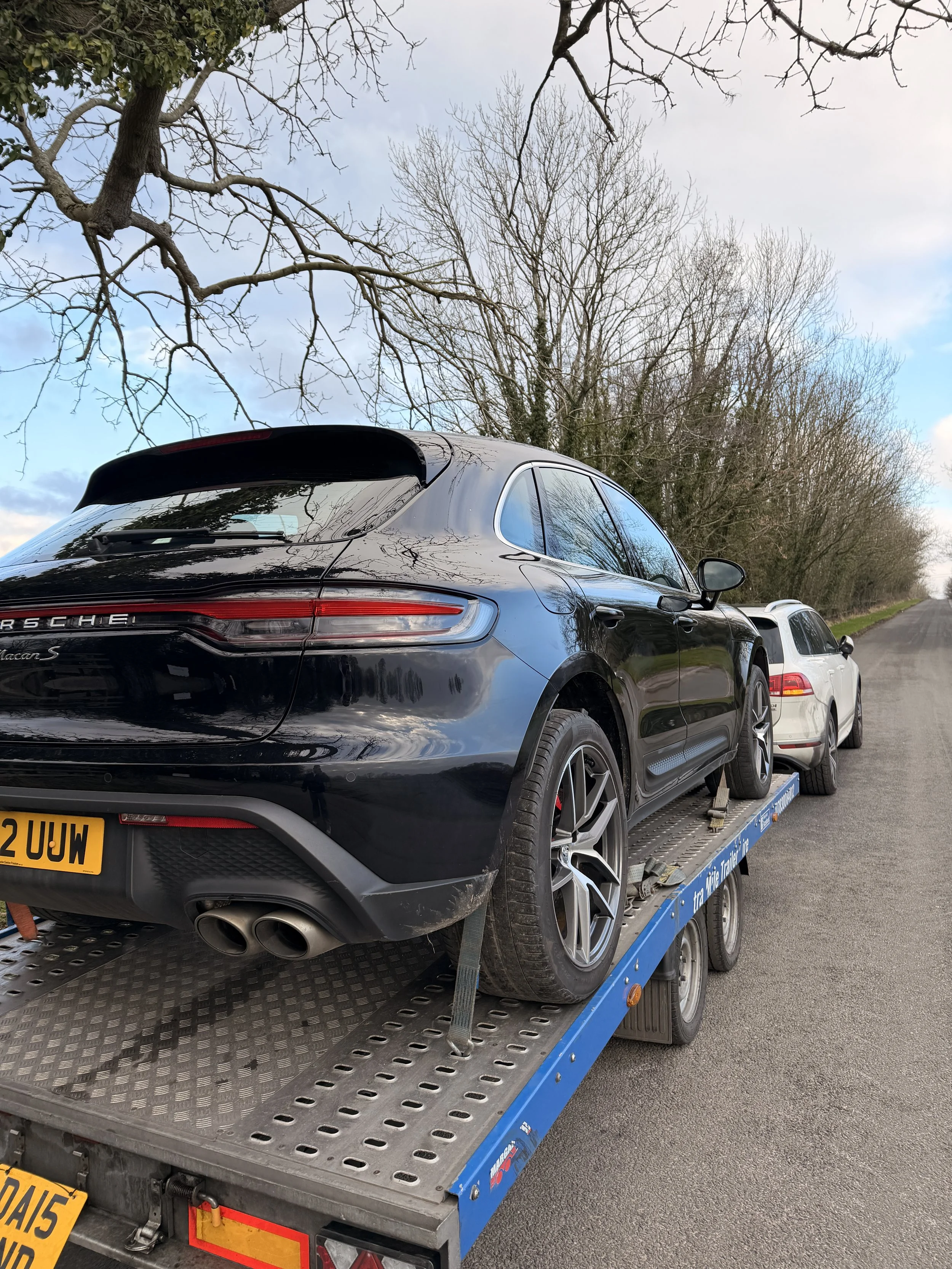 Black Porsche Macan S on a flatbed trailer with trees and a cloudy sky in the background.