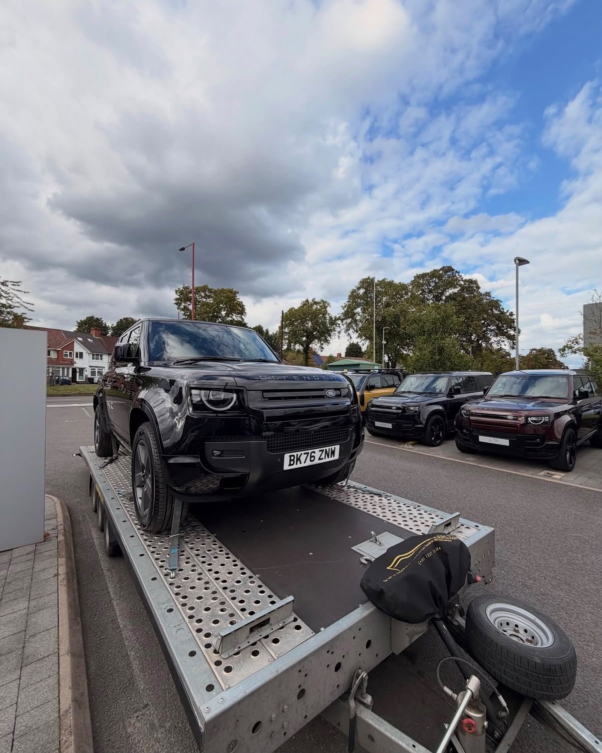 Black Land Rover Defender on a trailer, parked in a lot with other cars and trees in the background under a cloudy sky.