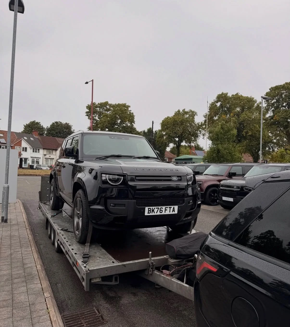 A black Land Rover Defender is being transported on a flatbed tow truck in a parking lot, with other parked cars and residential houses in the background, and overcast sky.