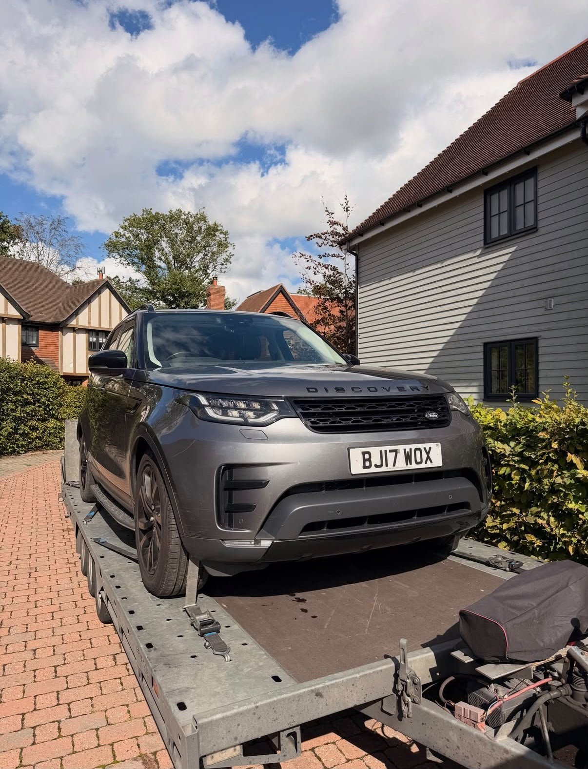 A grey Land Rover Discovery SUV on a flatbed trailer in a residential driveway, with houses, bushes, and trees in the background under a partly cloudy sky.