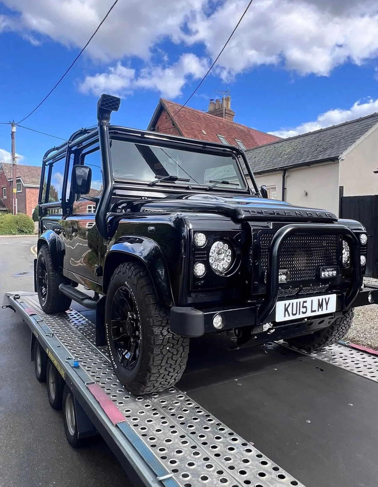 A black Land Rover Defender on a flatbed tow truck against a background of residential houses and a blue sky with clouds.