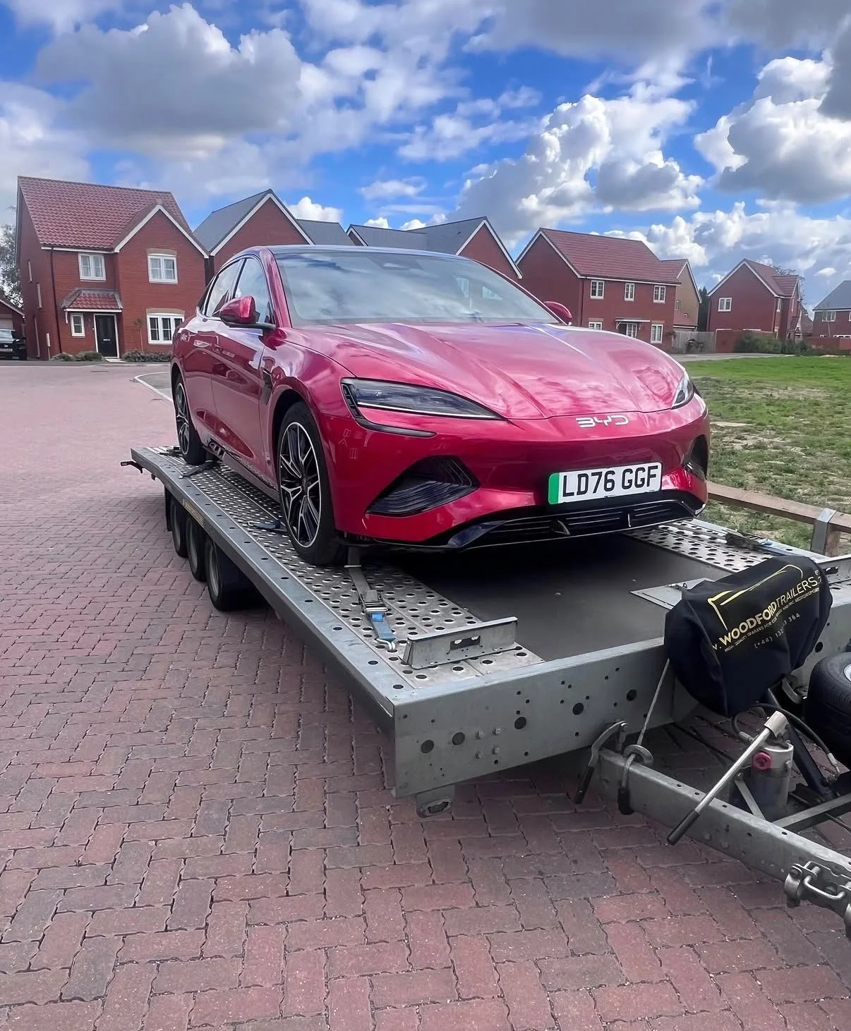 Red electric car on a silver trailer in a residential neighborhood with brick houses and a partly cloudy sky.