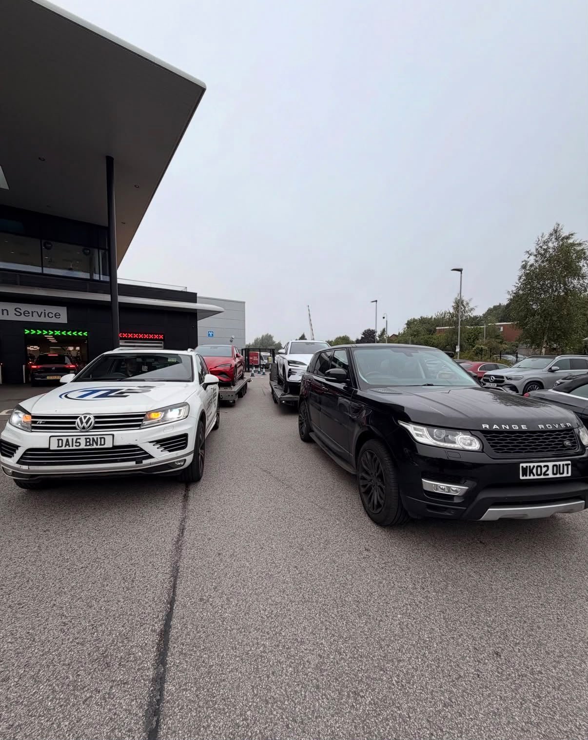 Several cars parked outside a building with a modern architectural design, including a Volkswagen, a Range Rover, and other vehicles. There is an overcast sky and some trees in the background.