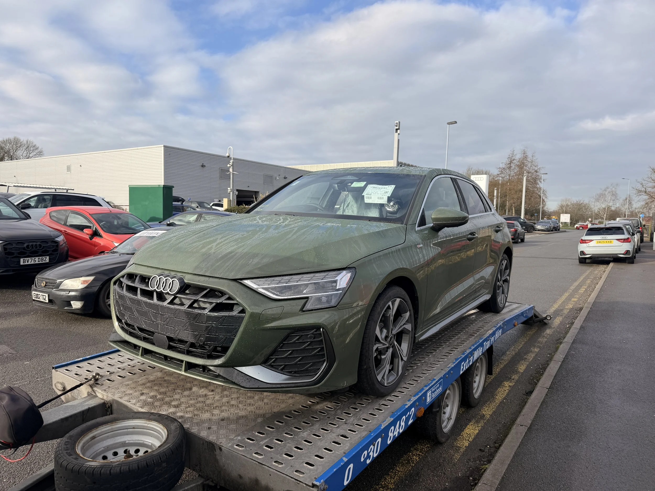 Green Audi car on a flatbed tow truck in a parking lot with other vehicles and a large white building with a blue sky and clouds in the background.