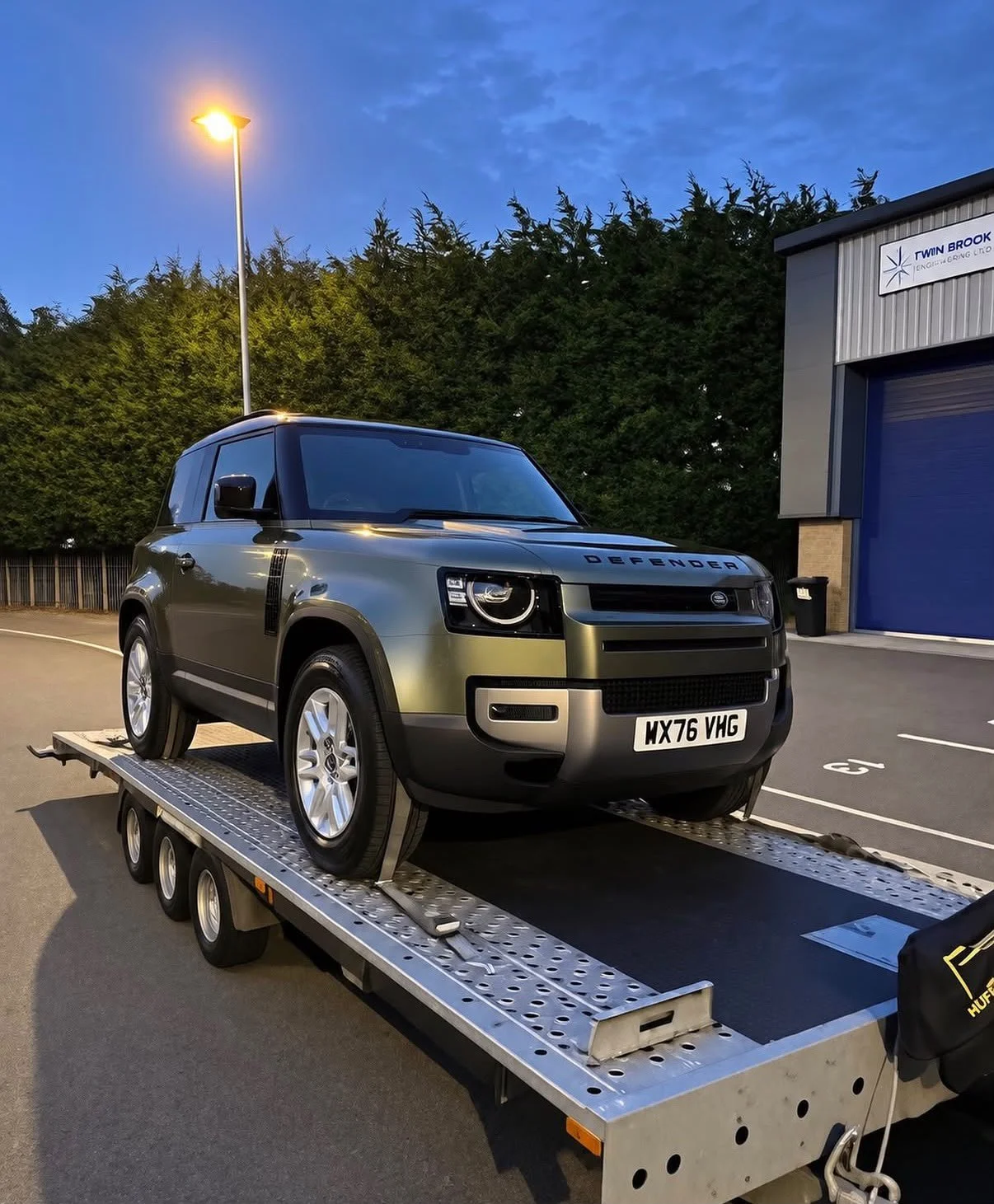 A black and gray Land Rover Defender on a flatbed tow truck in a parking lot during twilight, with a blue building and green trees in the background.