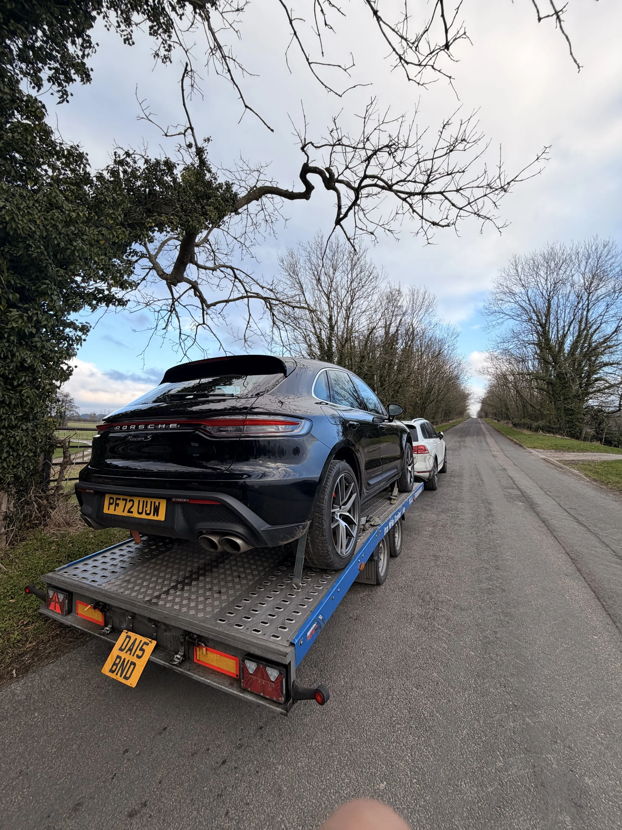 A black Porsche Macan S being transported on a flatbed trailer on a rural road with a row of leafless trees and green fields in the background.