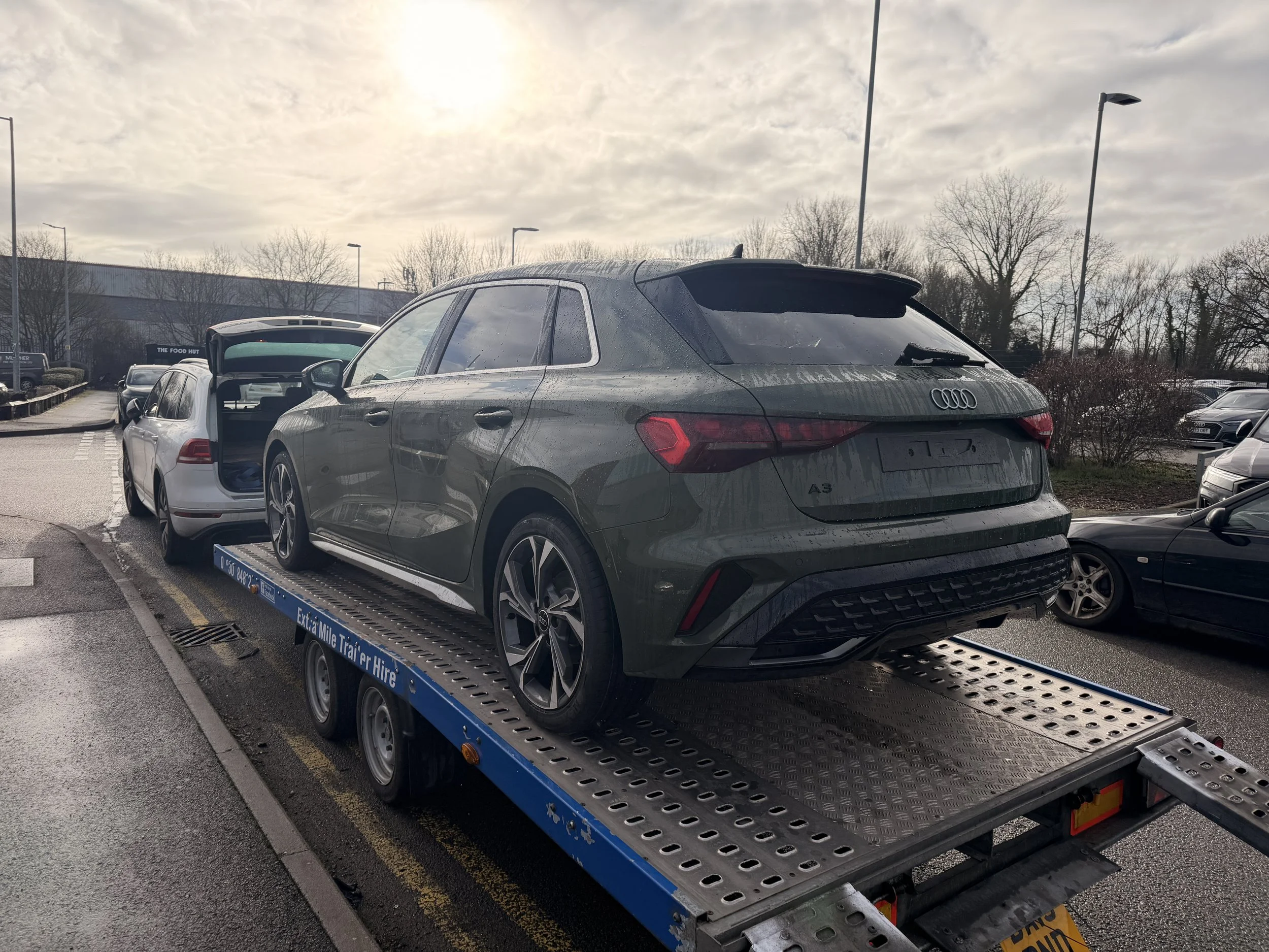 A silver Audi A3 car being transported on a flatbed tow truck in what appears to be a parking lot during daytime with cloudy skies.