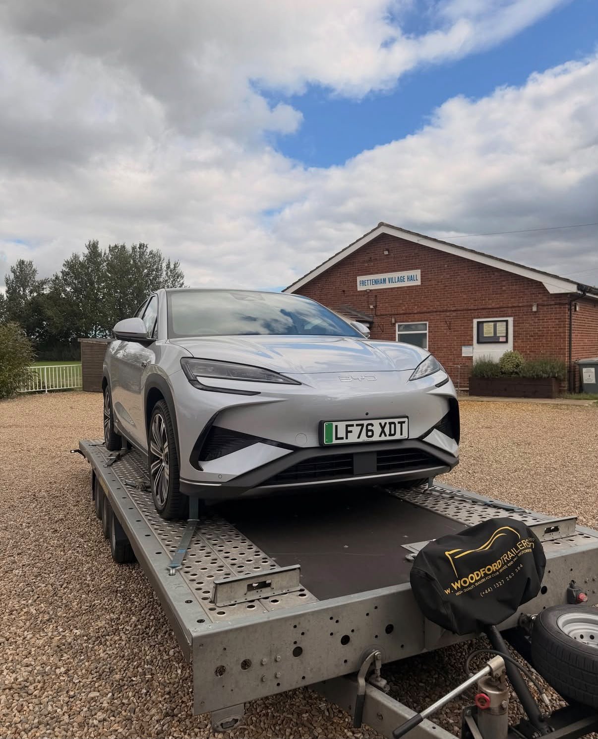 Silver electric car on a flatbed trailer in front of a brick village hall building, with a cloudy sky above.