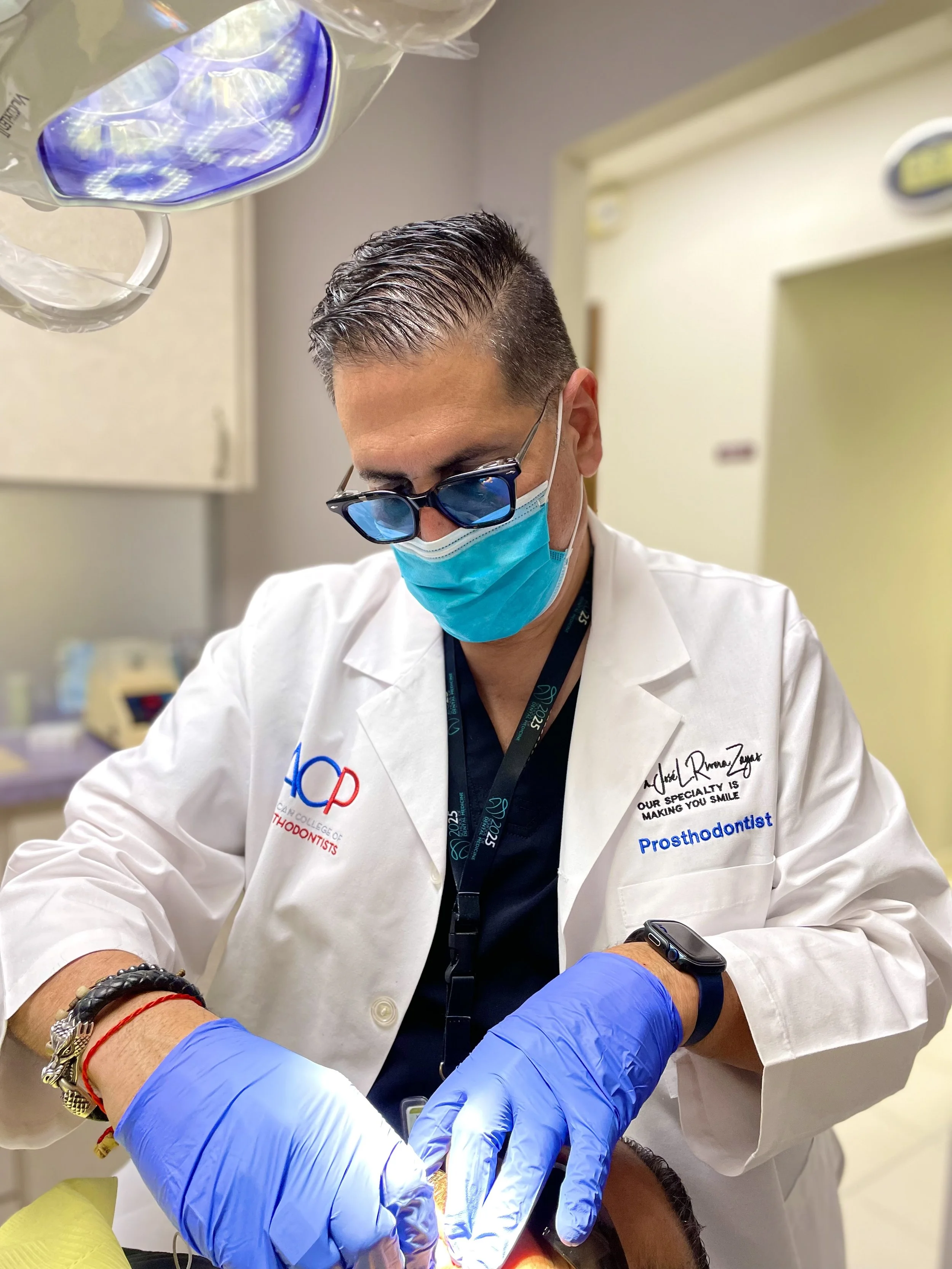Dr. Jose Rivera-Zayas wearing scrubs, a face mask, and protective glasses working on a patient's mouth in a dental clinic.