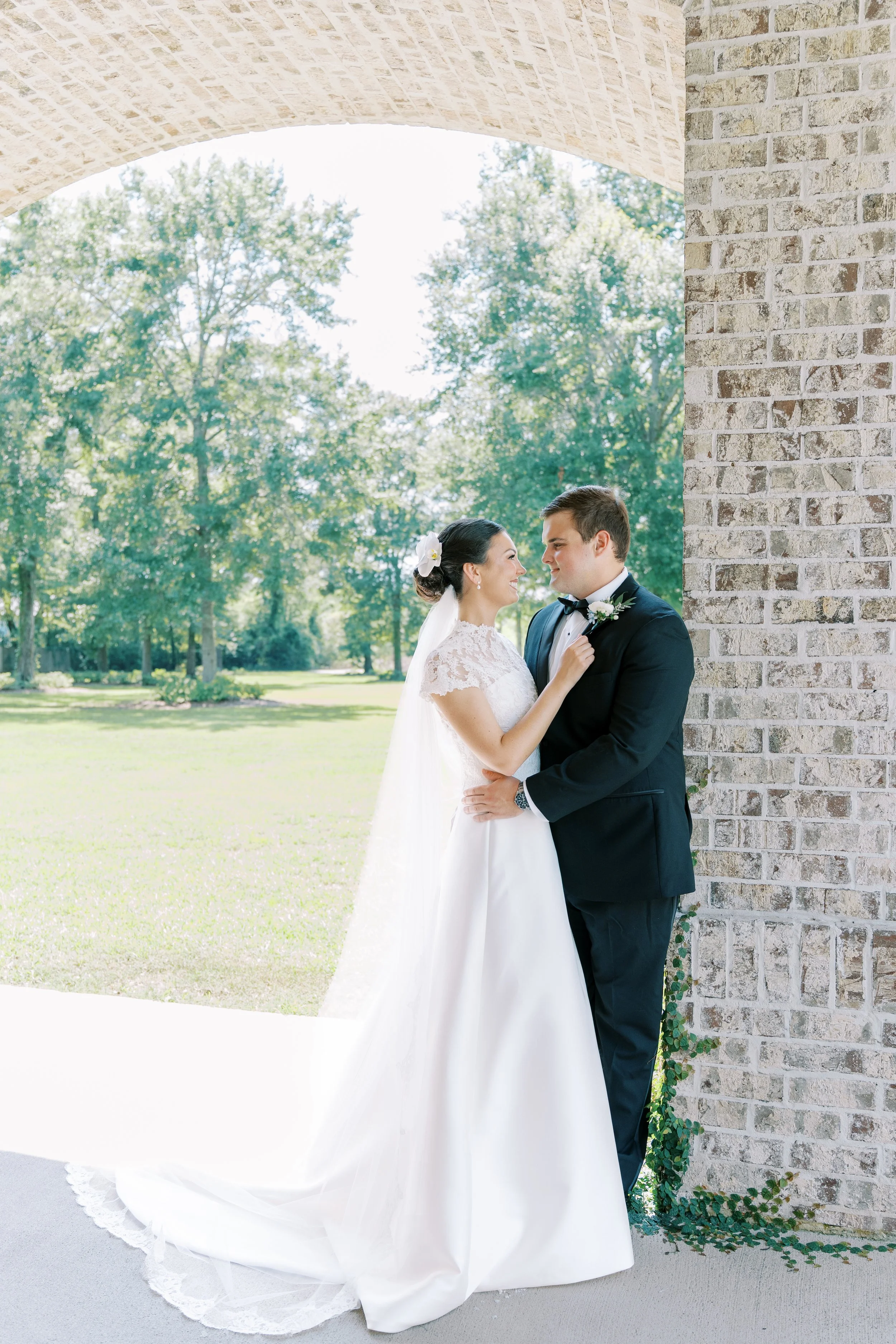 A bride and groom embrace under a brick archway, with trees and a sunny outdoor setting in the background.