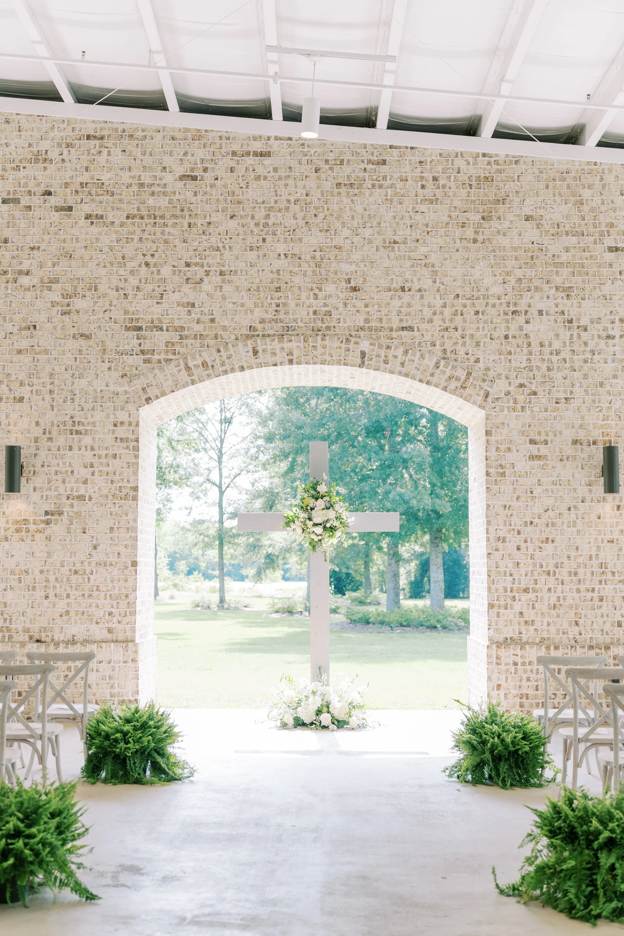 Interior of a chapel with a white cross adorned with flowers, set in front of a large window showing a green outdoor landscape.