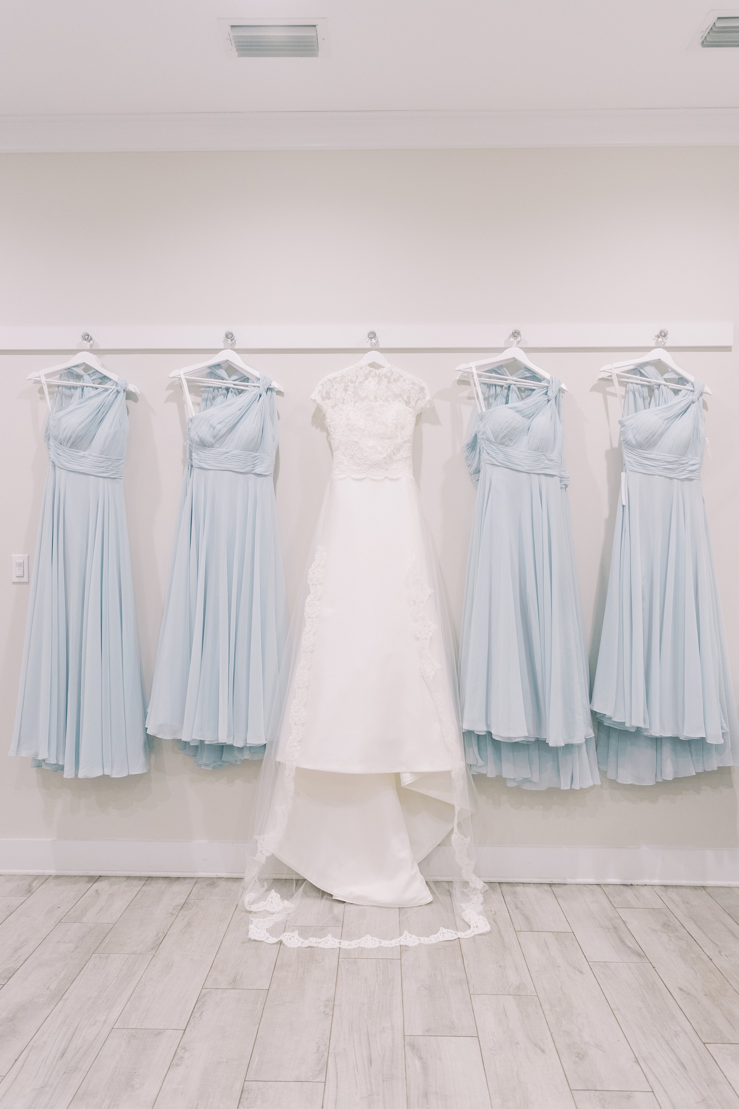 A white wedding dress with lace details on a hanger, placed in front of five light blue bridesmaid dresses hanging on a rack against a white wall.