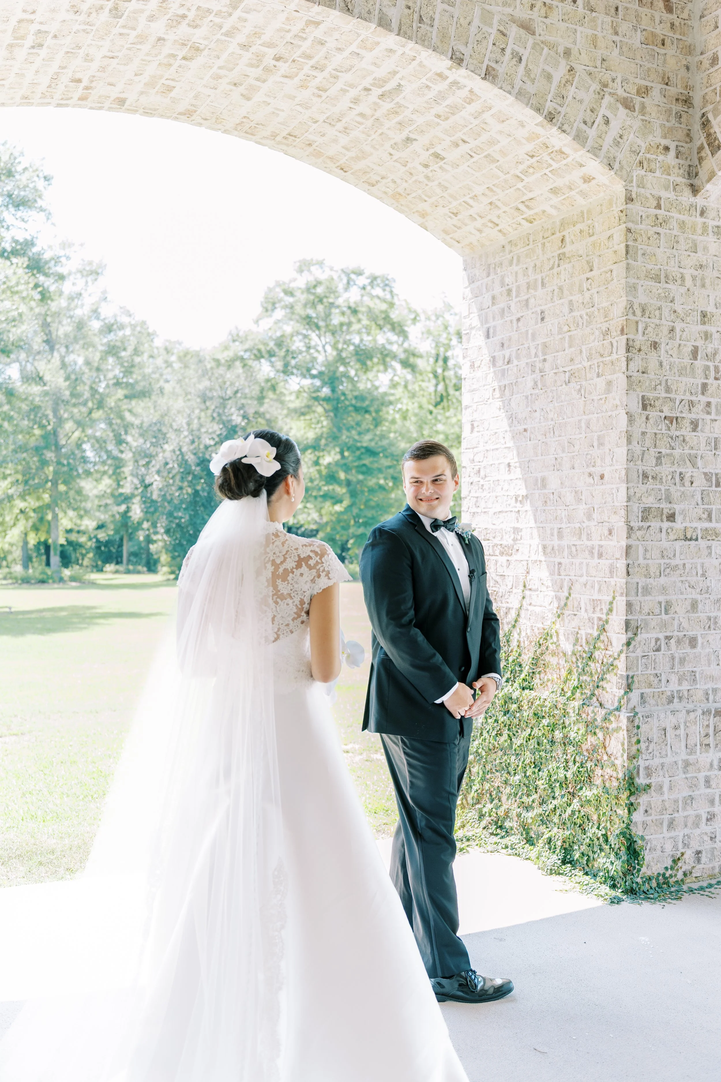 A bride and groom stand together under a large stone archway outdoors, with trees and grass in the background on a sunny day.