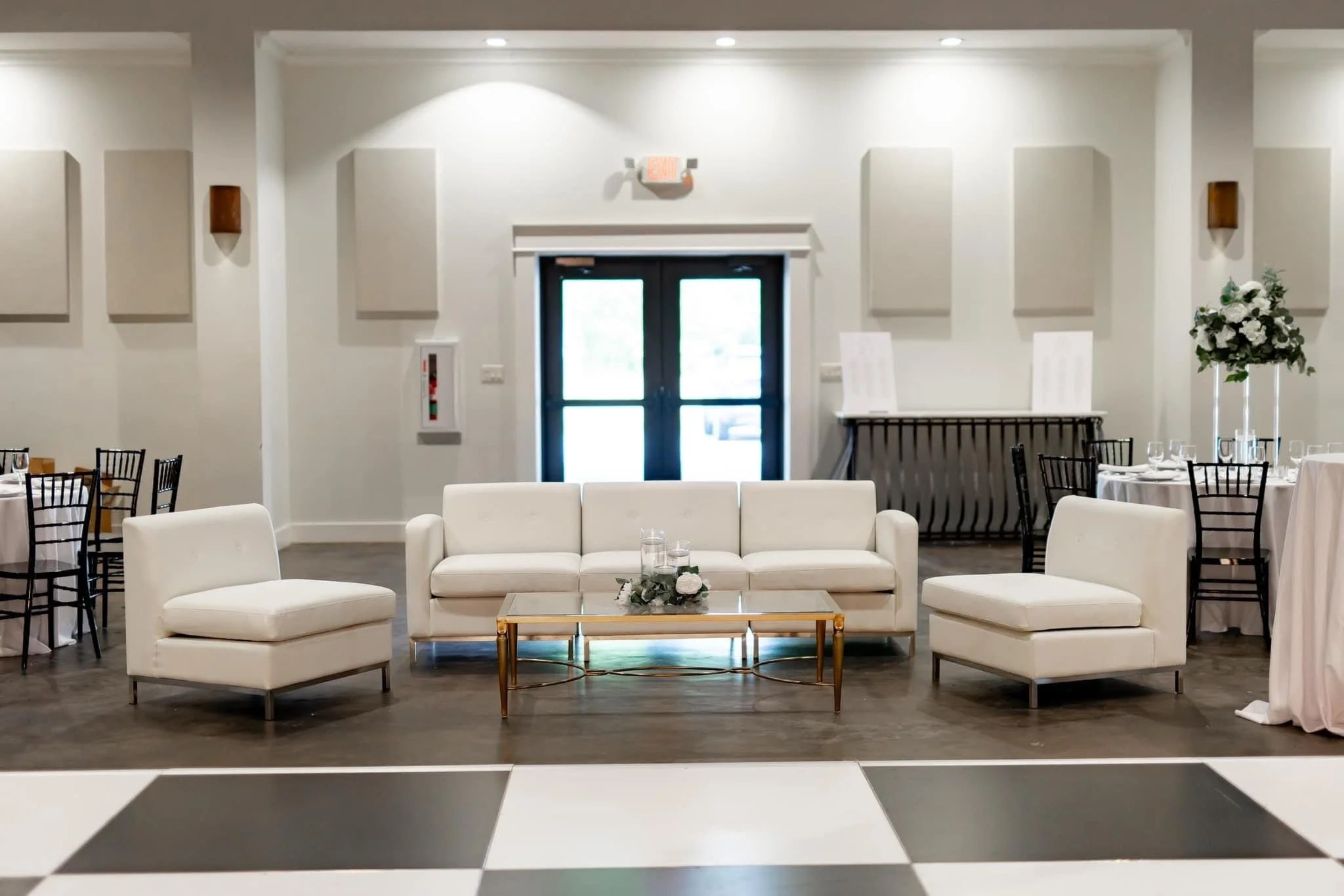 Elegant wedding or event reception area with white couches and chairs, a glass-top coffee table, floral centerpiece, and tables set with glassware, in a spacious, well-lit room.