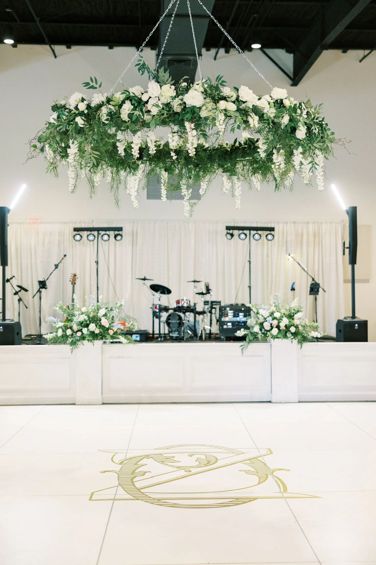 Wedding reception stage with a floral chandelier overhead, white floral arrangements on the stage, and musical instruments set up for a performance.