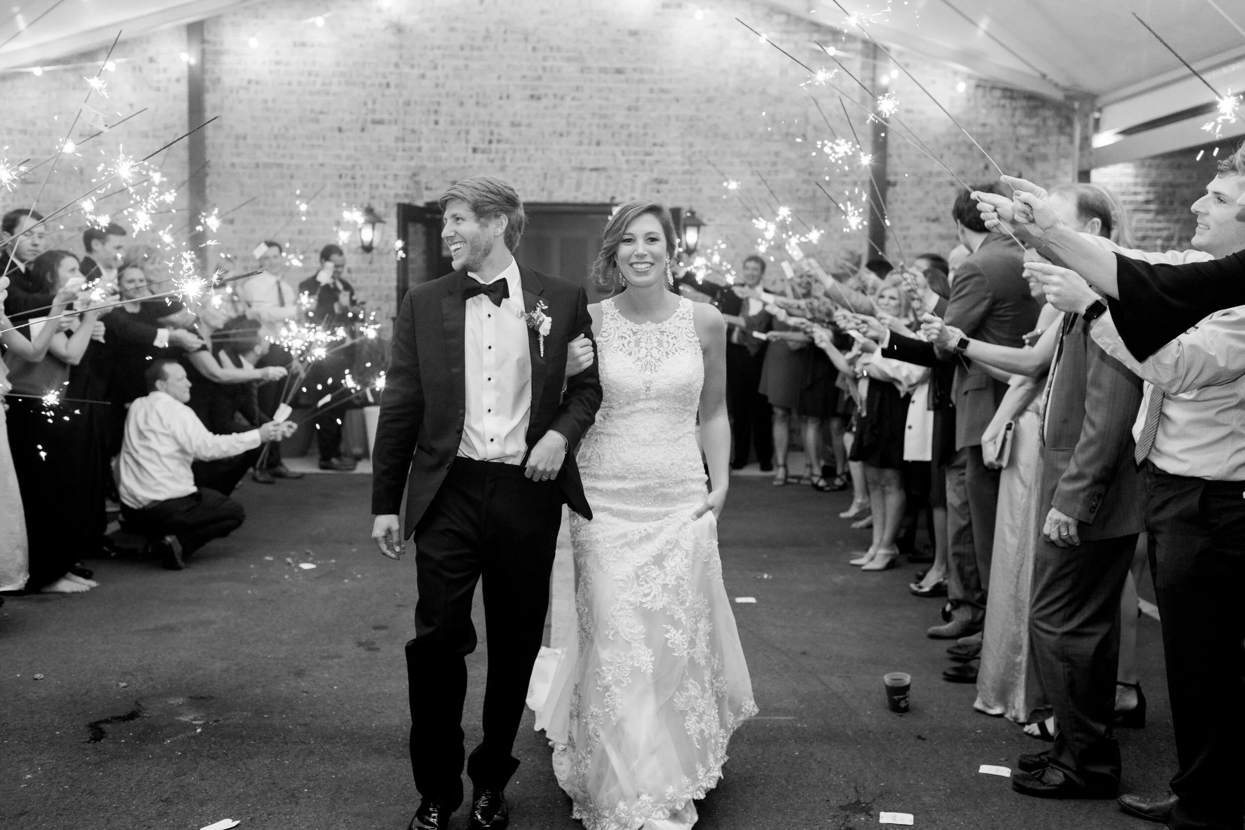 A black and white photo of a newlywed couple walking through a crowd of friends and family holding sparklers during a celebration.
