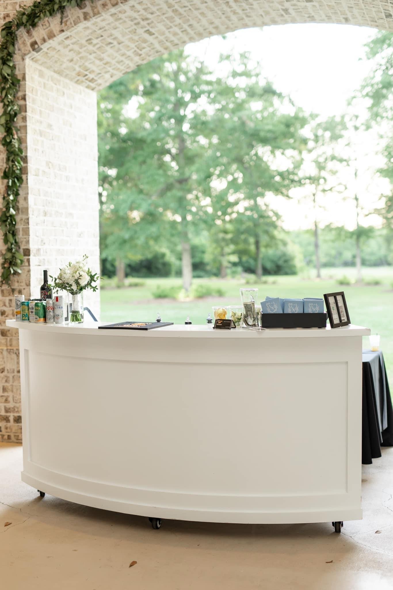 White bar counter with drinks, flowers, and event items, set in an outdoor pavilion with green trees in the background.