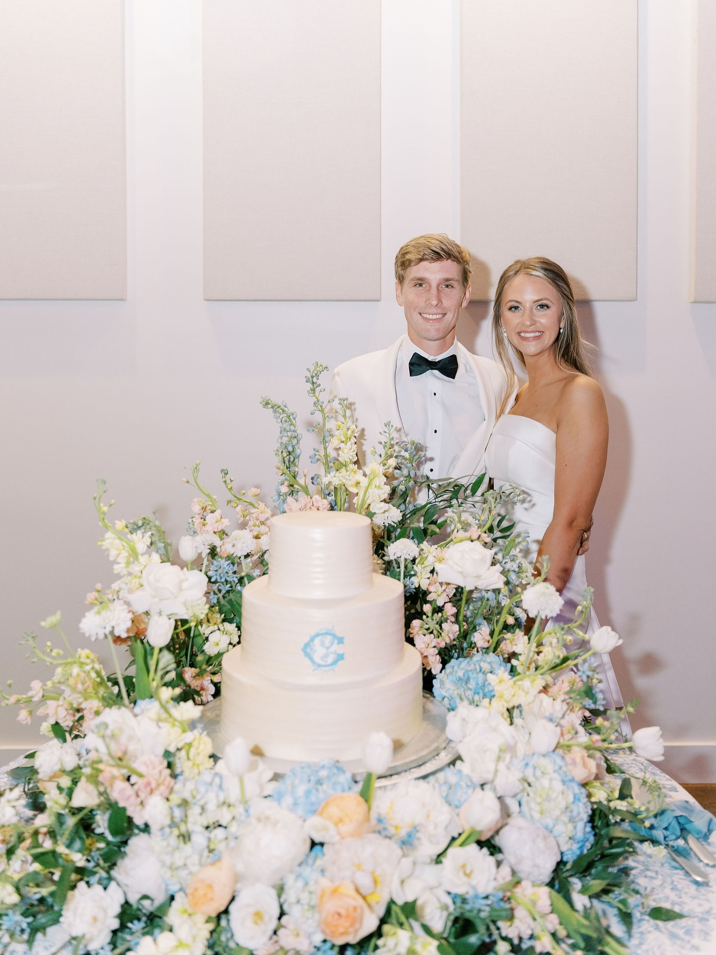 A newlywed couple standing behind a wedding cake decorated with flowers at their wedding reception.