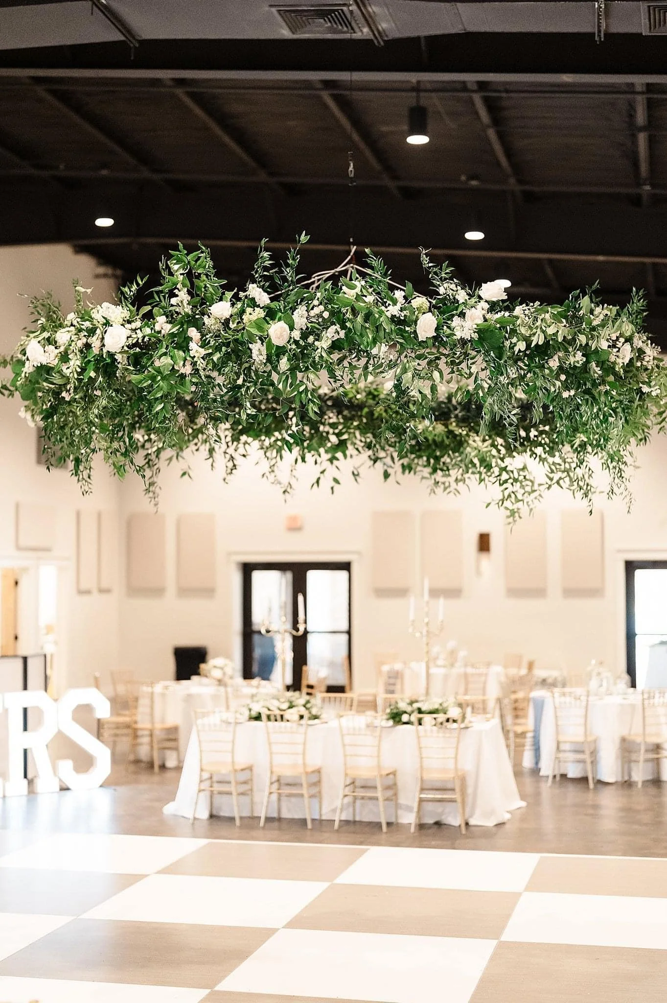 A large hanging floral arrangement with green foliage and white flowers above a decorated banquet hall with round tables, white tablecloths, gold chairs, and candelabras.