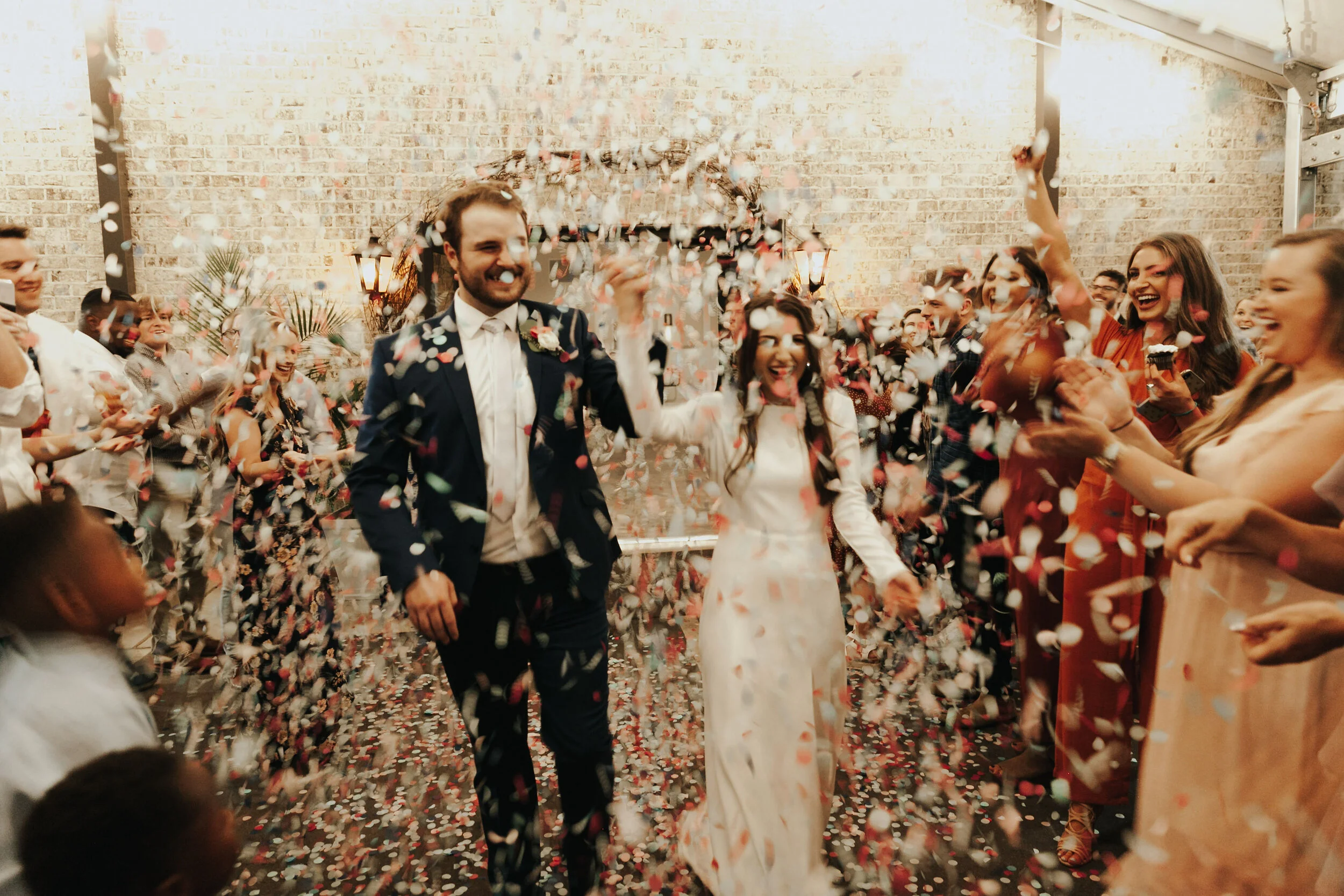 A newlywed couple walks through a shower of colorful confetti at their wedding reception, surrounded by joyful guests in an indoor setting with brick walls.