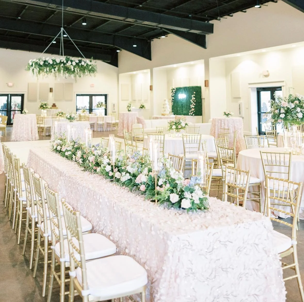 Decorated wedding reception hall with round tables, floral centerpieces, a long sweetheart table with flowers, gold chairs, and a cake table in the background.