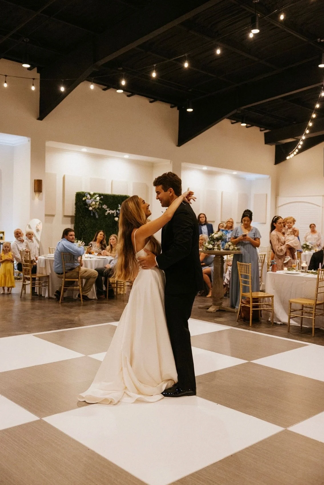 A bride and groom dance in the center of a wedding reception hall with guests seated at tables around them.