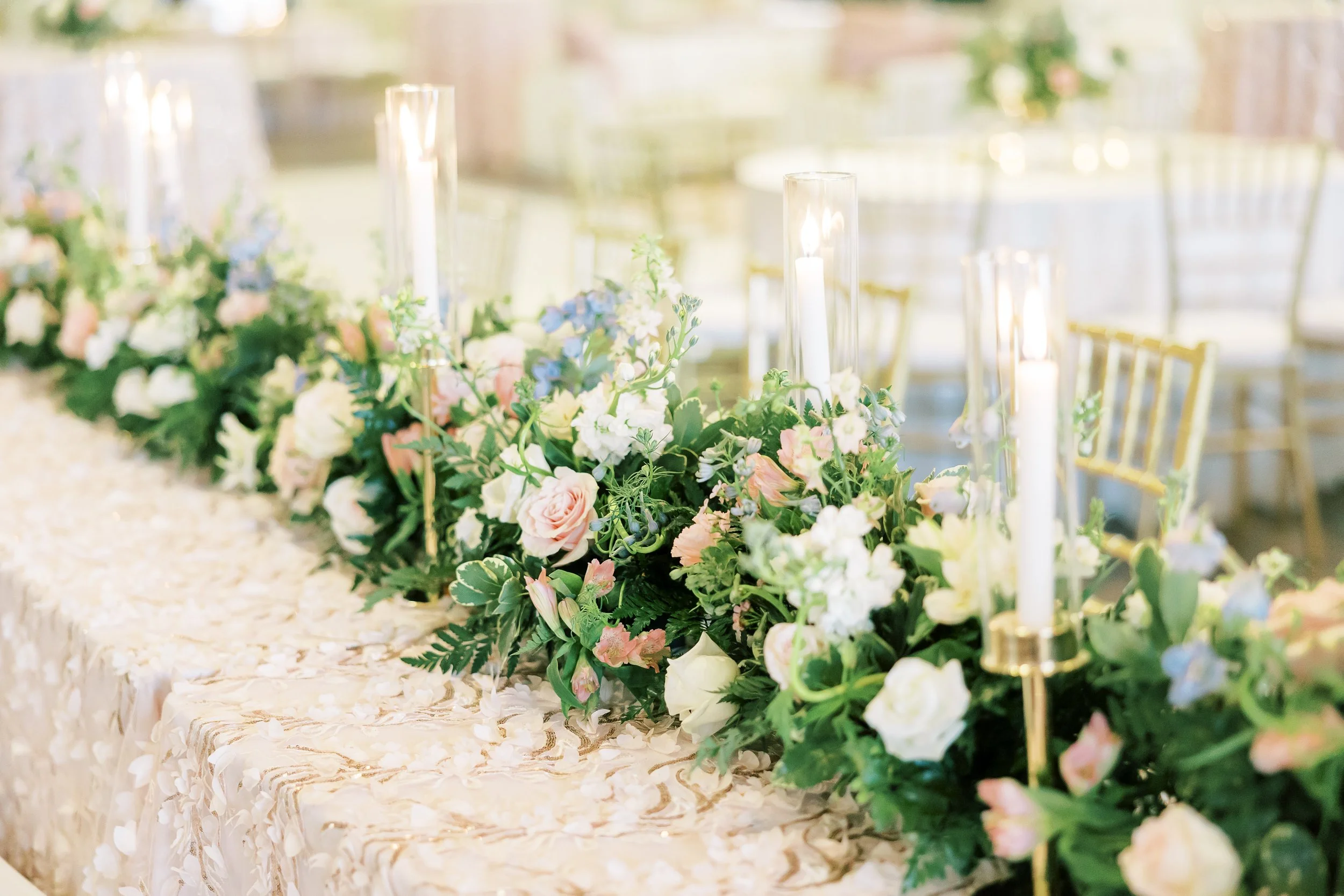 Elegant wedding or event table decorated with a long floral arrangement of pink, white, and blue flowers, surrounded by candles in tall glass holders, with gold chairs in the background.