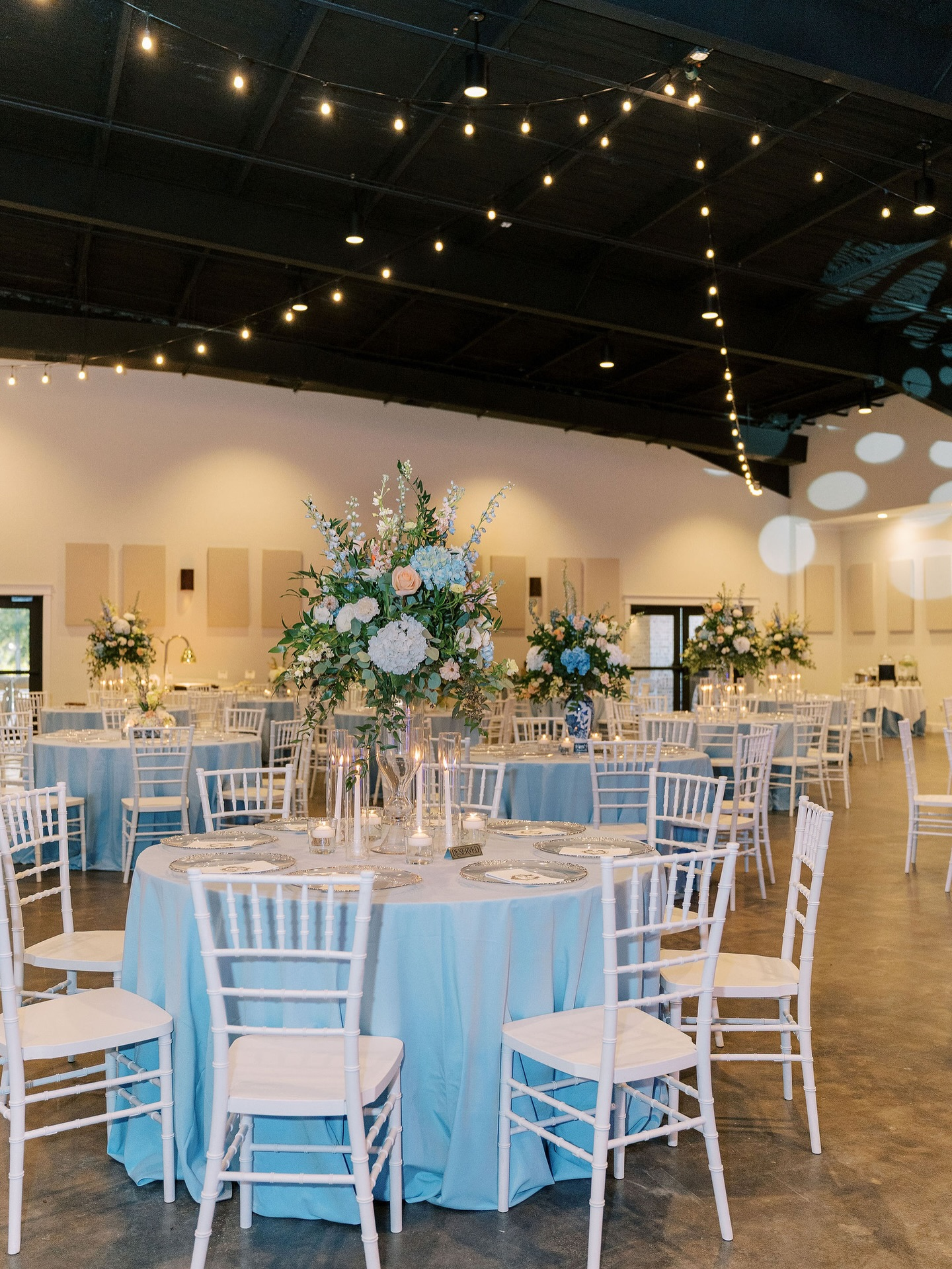 Elegant event space decorated for a wedding reception with round tables draped in light blue tablecloths, each set with plates and silverware. Tall floral centerpieces featuring white and blush flowers with greenery are on each table. White chairs su