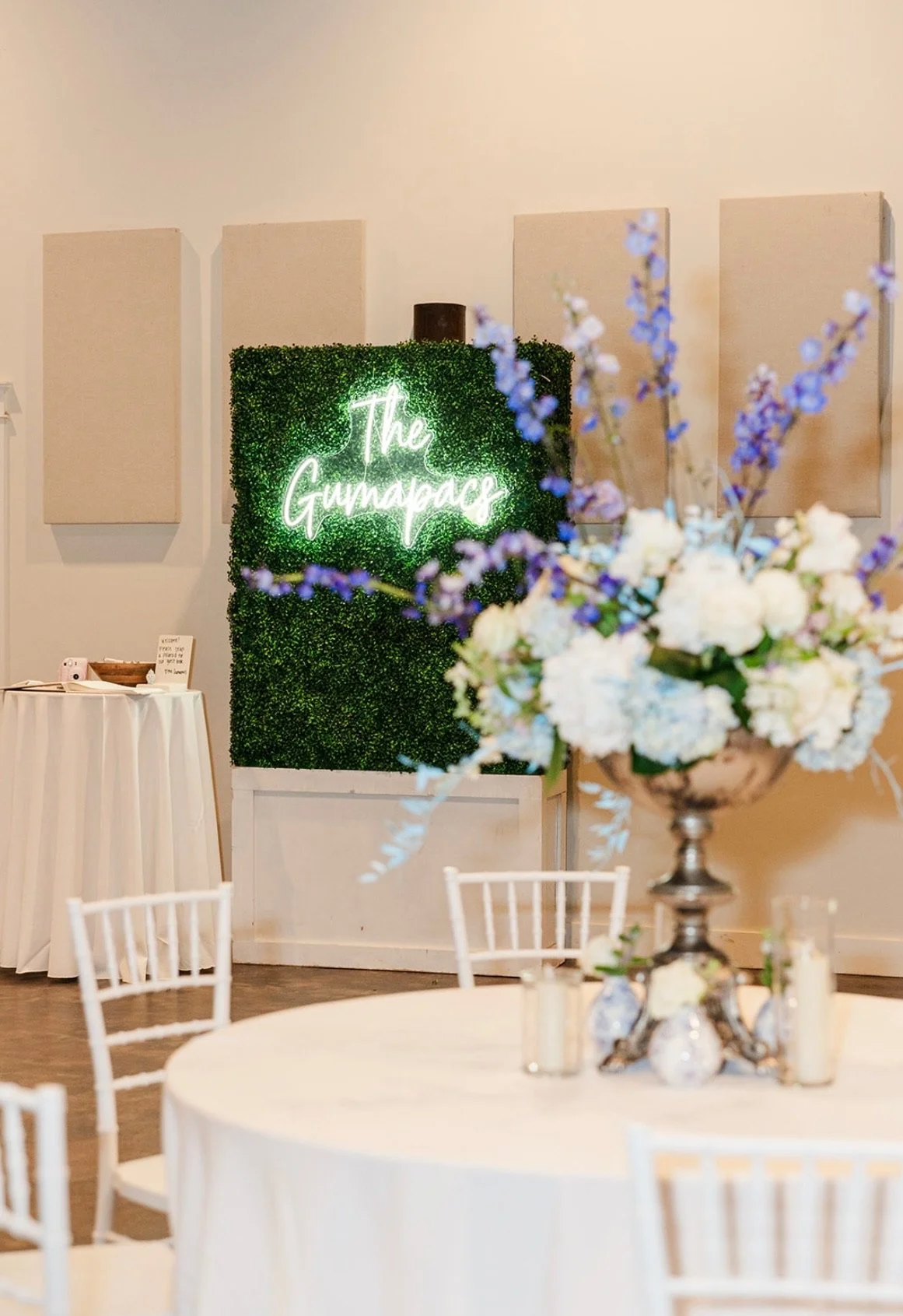 Neon sign reading 'The Gumdrop' on a green hedge wall, with floral arrangements and round tables in the foreground.
