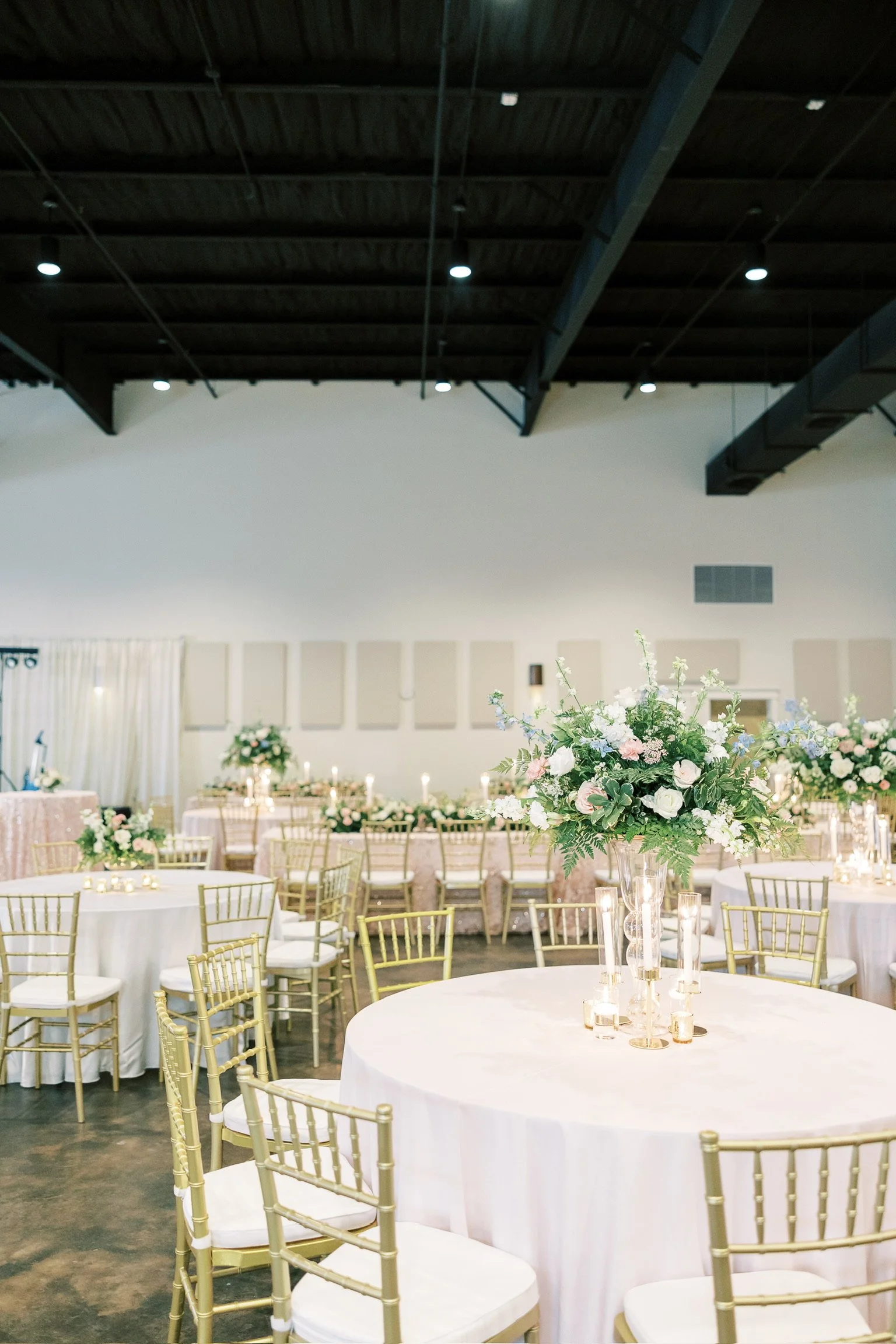 Elegant event space decorated with gold chairs, white tablecloths, and floral centerpieces with candles.