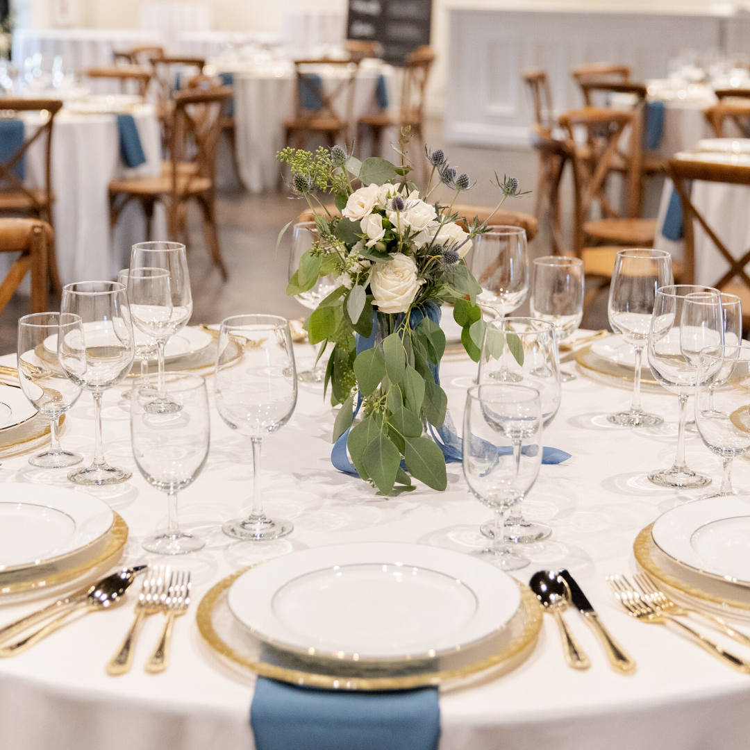 Elegant banquet table setup with white tablecloth, gold-rimmed plates, gold cutlery, multiple wine glasses, and a floral centerpiece with white flowers and green leaves in a venue with wooden chairs and blue napkins.