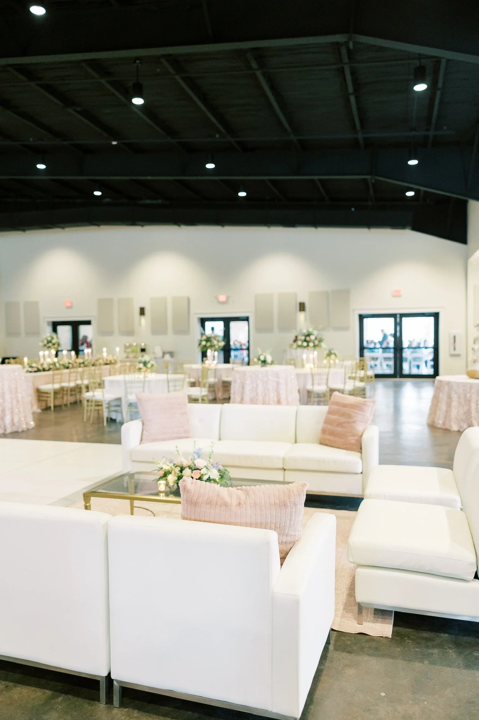 A spacious event hall decorated with white and blush-colored furniture, floral arrangements, and round tables covered with blush tablecloths, set up for a celebration or wedding reception.