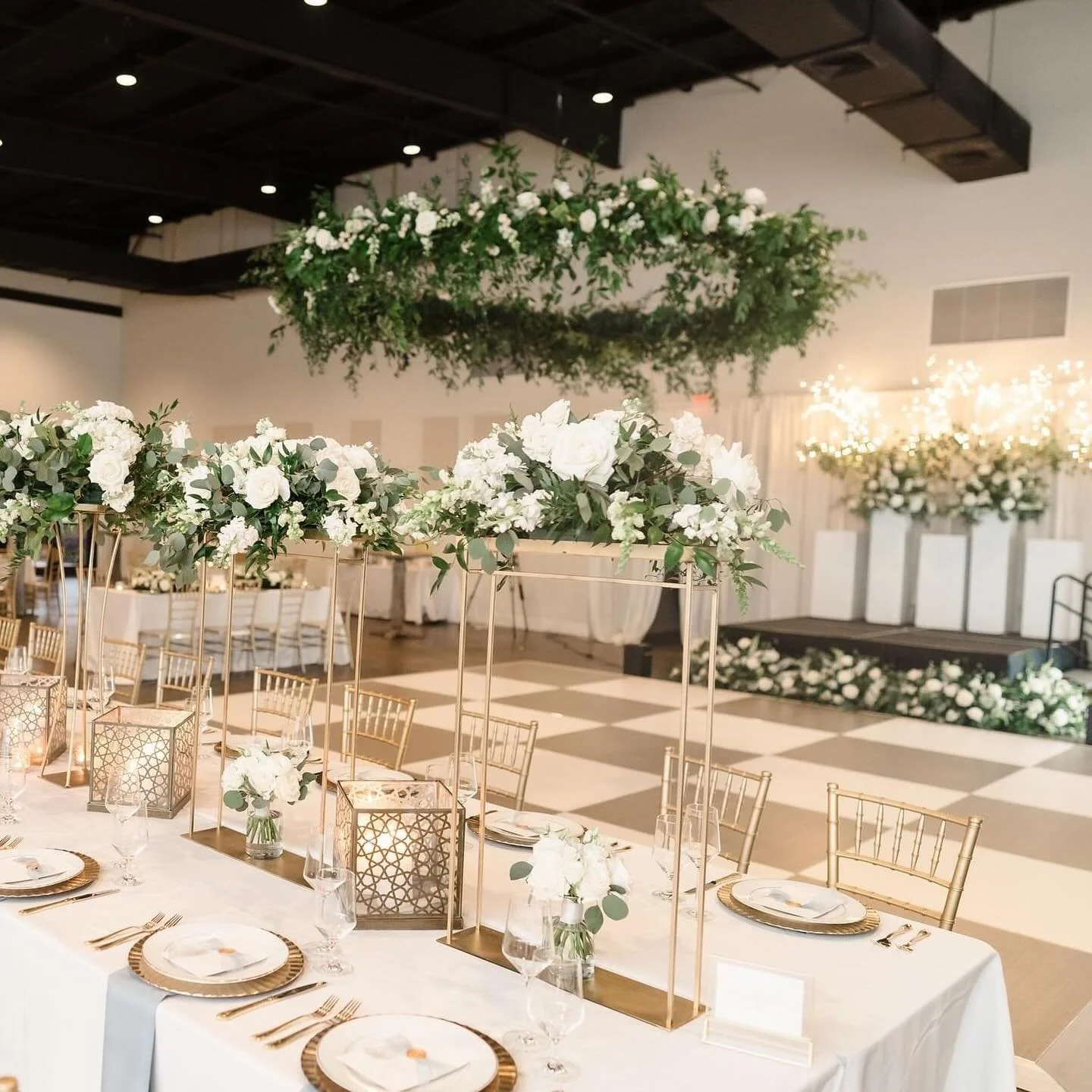 Elegant wedding reception setup with white floral centerpieces, gold chairs, and a stage decorated with white flowers and greenery.