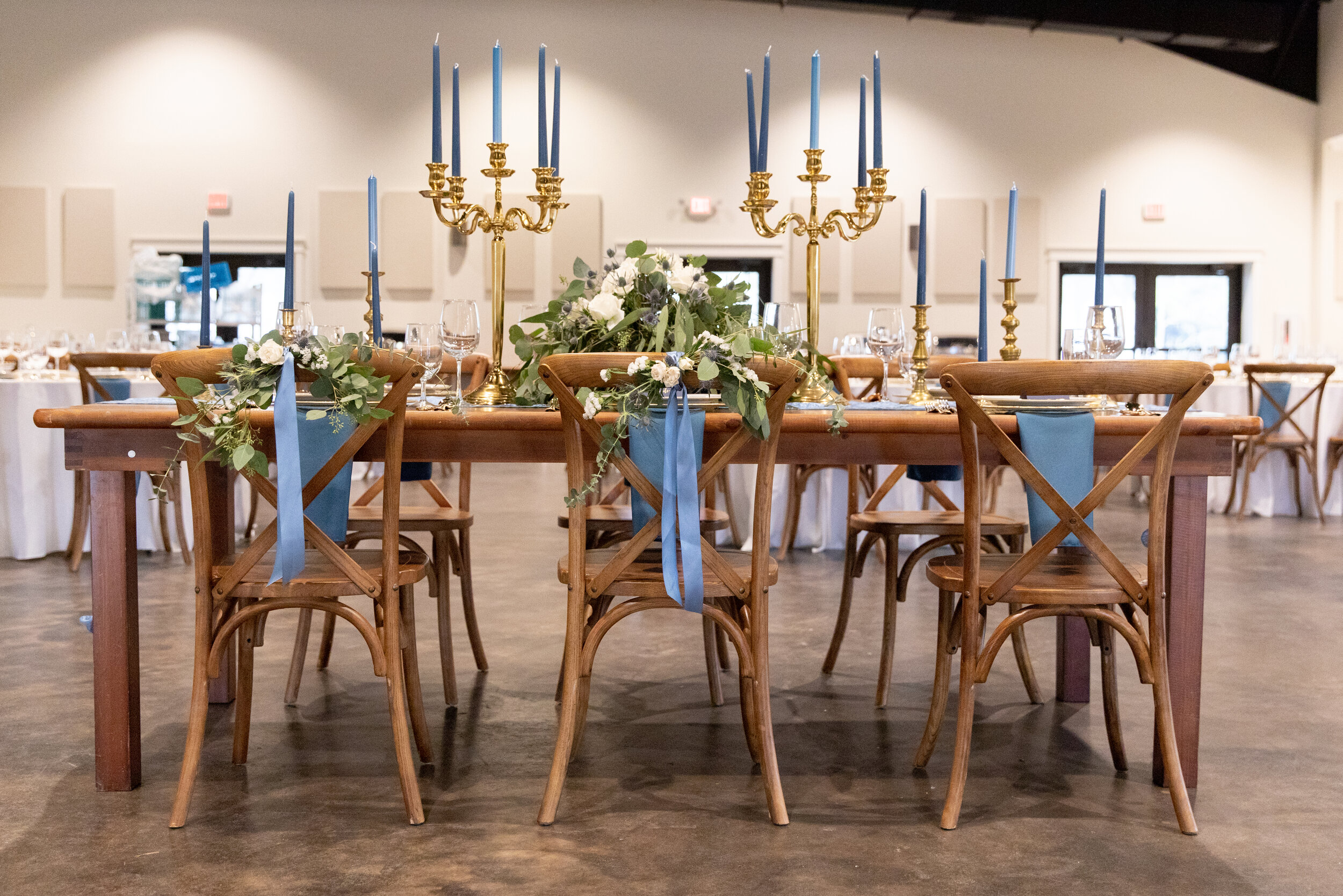 A decorated banquet table with blue candles, gold candelabras, floral arrangements, and blue ribbons in a banquet hall.