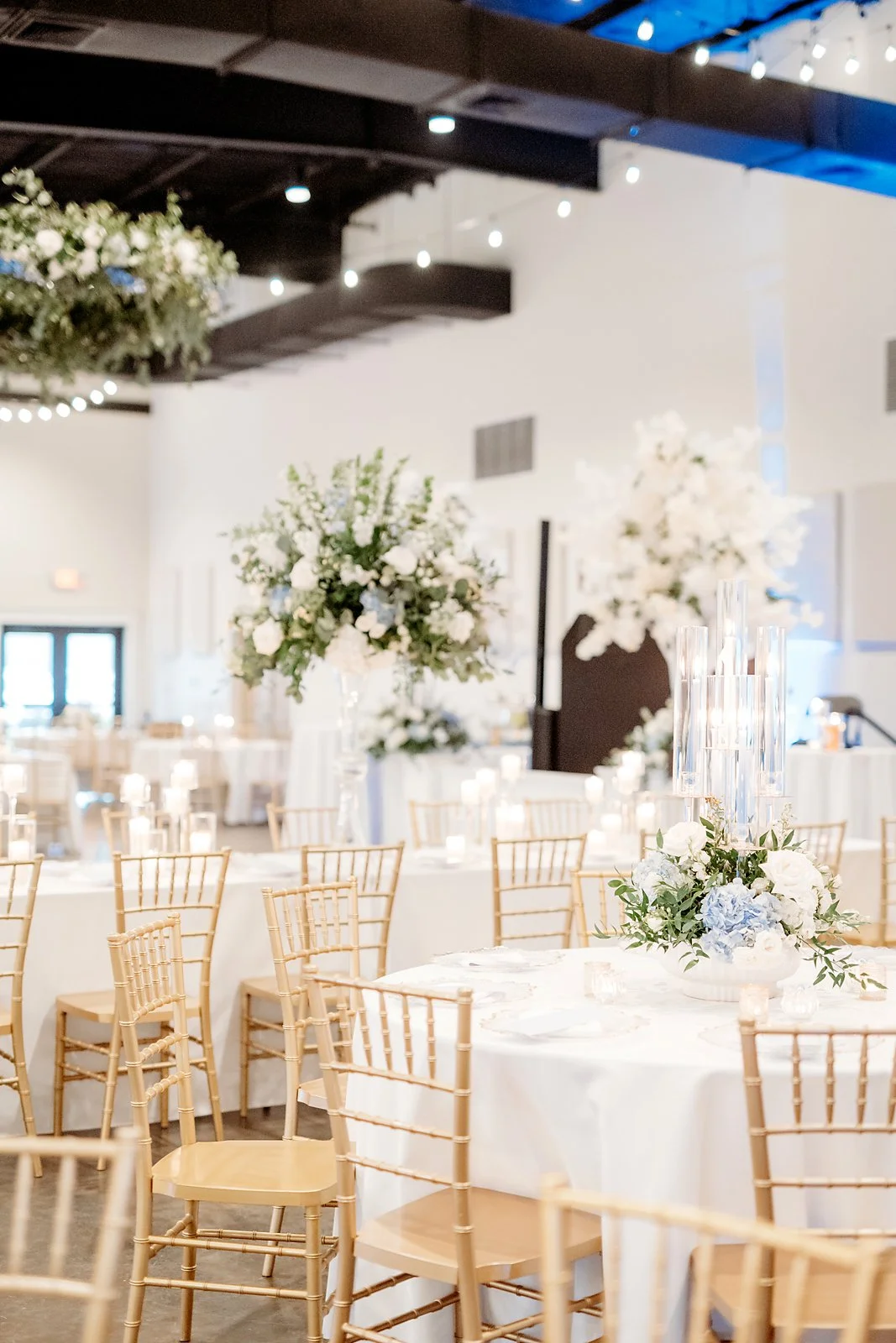 Elegant event space decorated with white flowers and tall glass candle holders, set for a wedding reception with round tables, gold chairs, and string lights overhead.