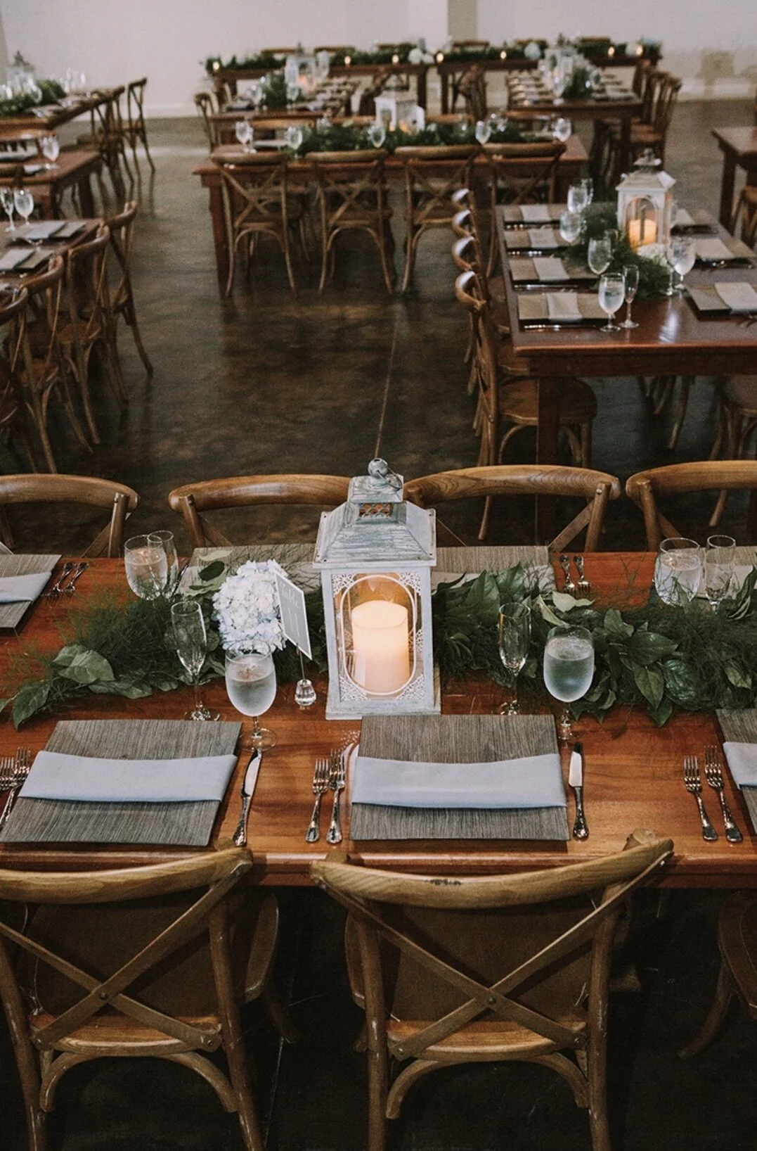 Decorated banquet table with a white lantern containing a candle, greenery, white flowers, glasses of water, silverware, and folded napkins, set for a formal event or reception.
