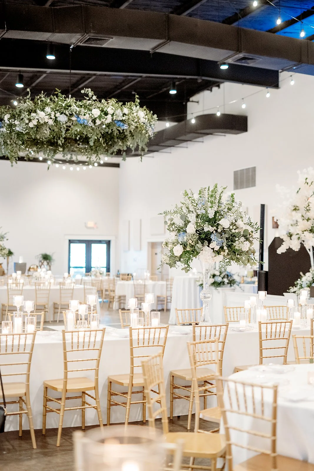 Elegant event space decorated with large floral arrangements on tables and hanging floral wreaths, white tablecloths, candles, and wooden chairs, with string lights overhead.