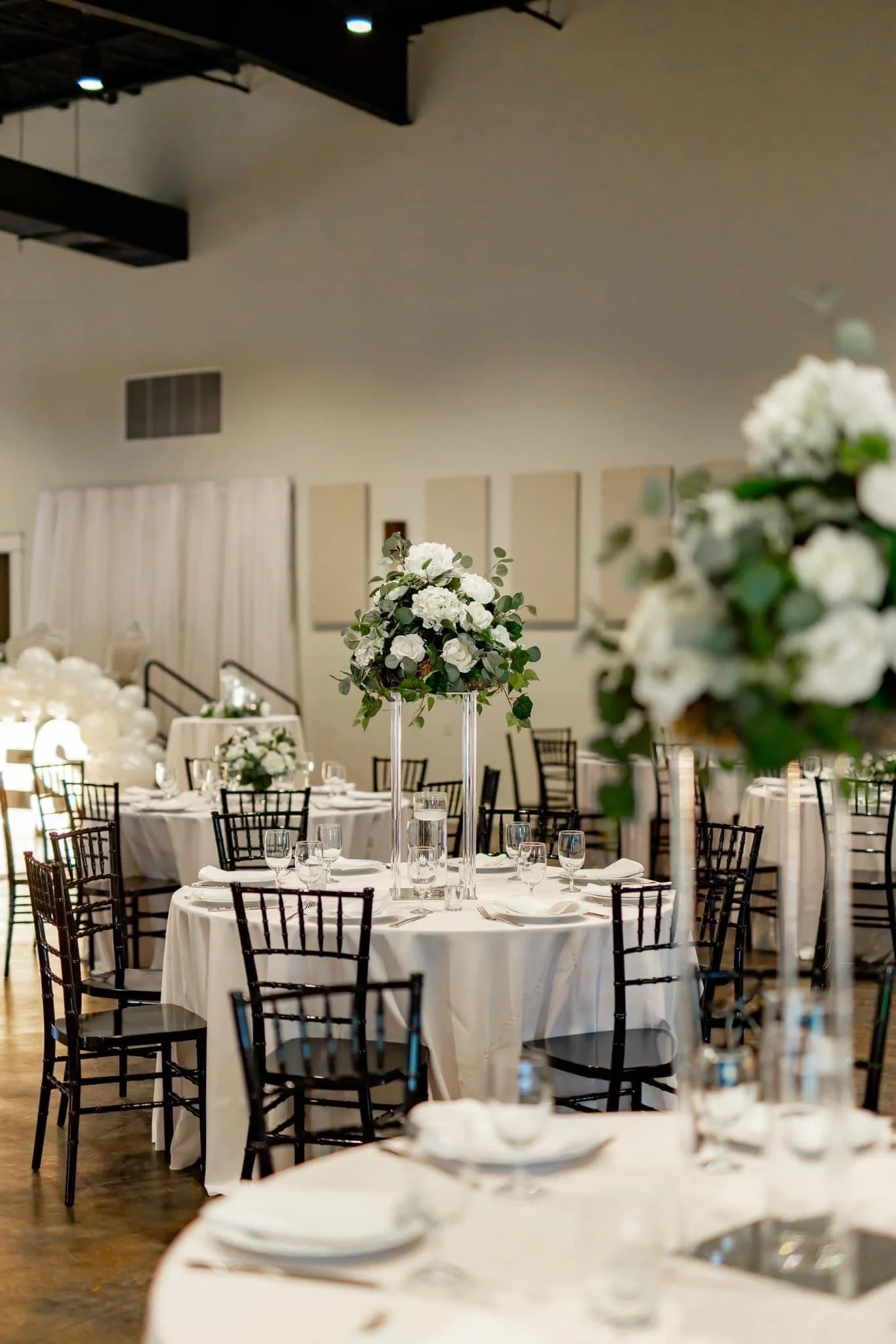 Elegant banquet hall decorated with white flowers on tall centerpieces, set with white tablecloths, glassware, and black chairs for a formal event.